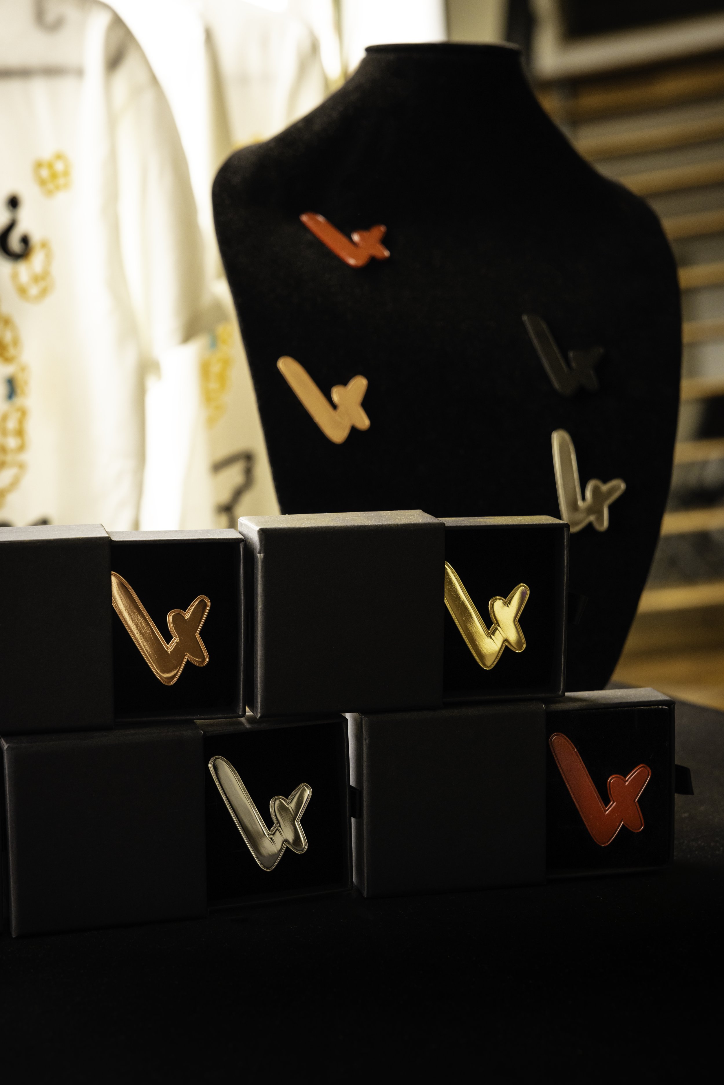 Black jewelry display bust with colorful letter-shaped pins attached, positioned in front of a display of black boxes with gold and red letter pins. The background has a white curtain with yellow and black patterns.