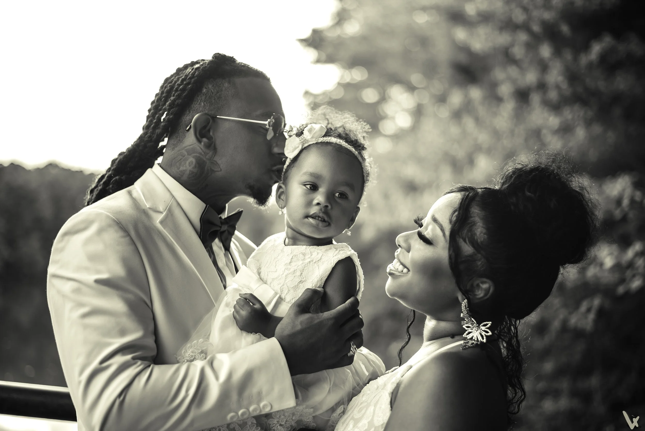 An African-American man in a tuxedo holding a young girl in a white dress, with a woman and another young girl close by, outdoors during daytime, in black and white.