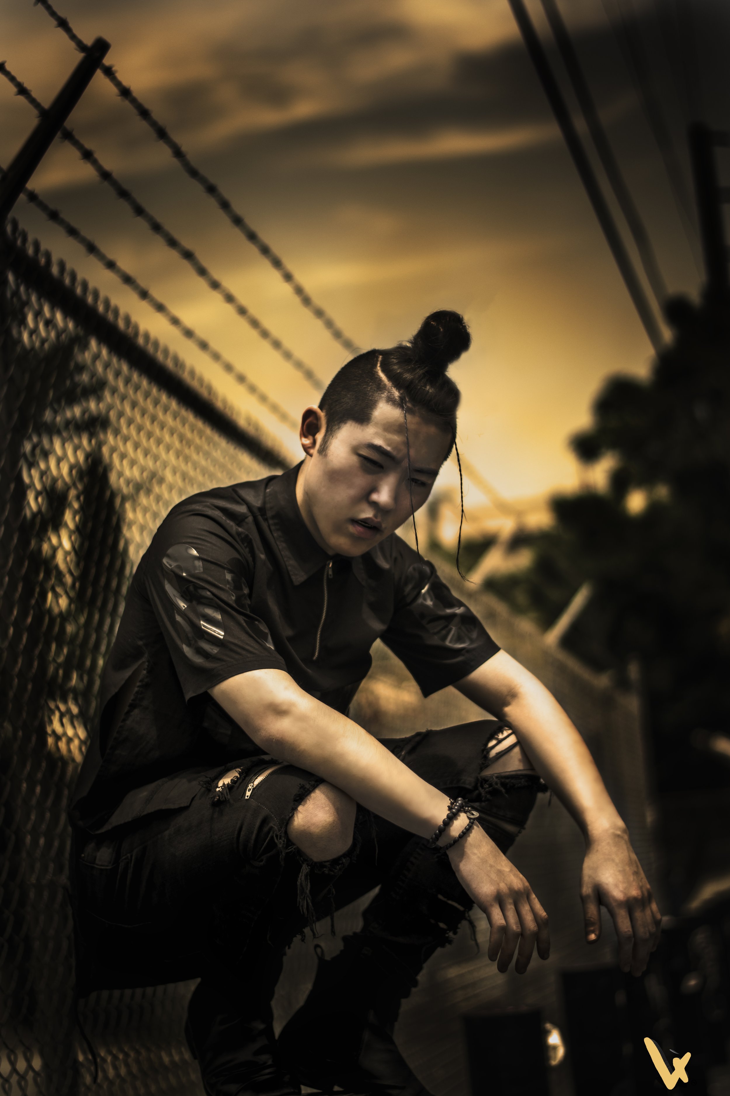 A young man in dark clothing sits on a ground near a chain-link fence during sunset, with a contemplative expression, arms resting on knees, and hands loosely clasped, against a cloudy sky and outdoor setting.