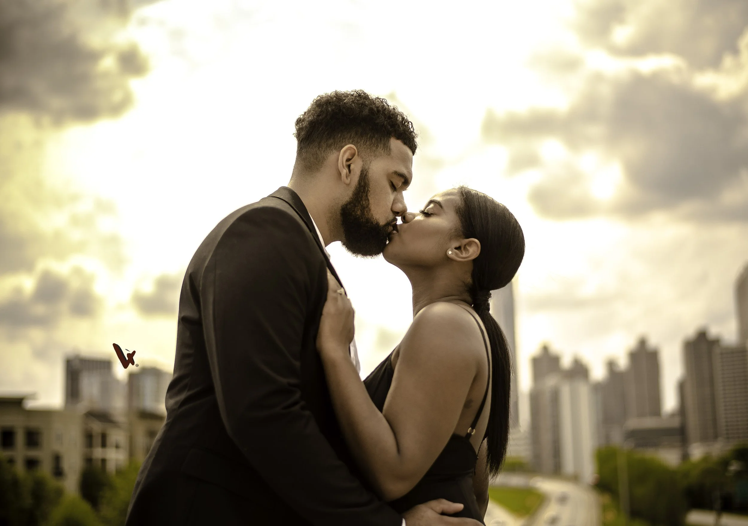 A man and woman sharing a kiss outdoors during sunset with city buildings in the background.