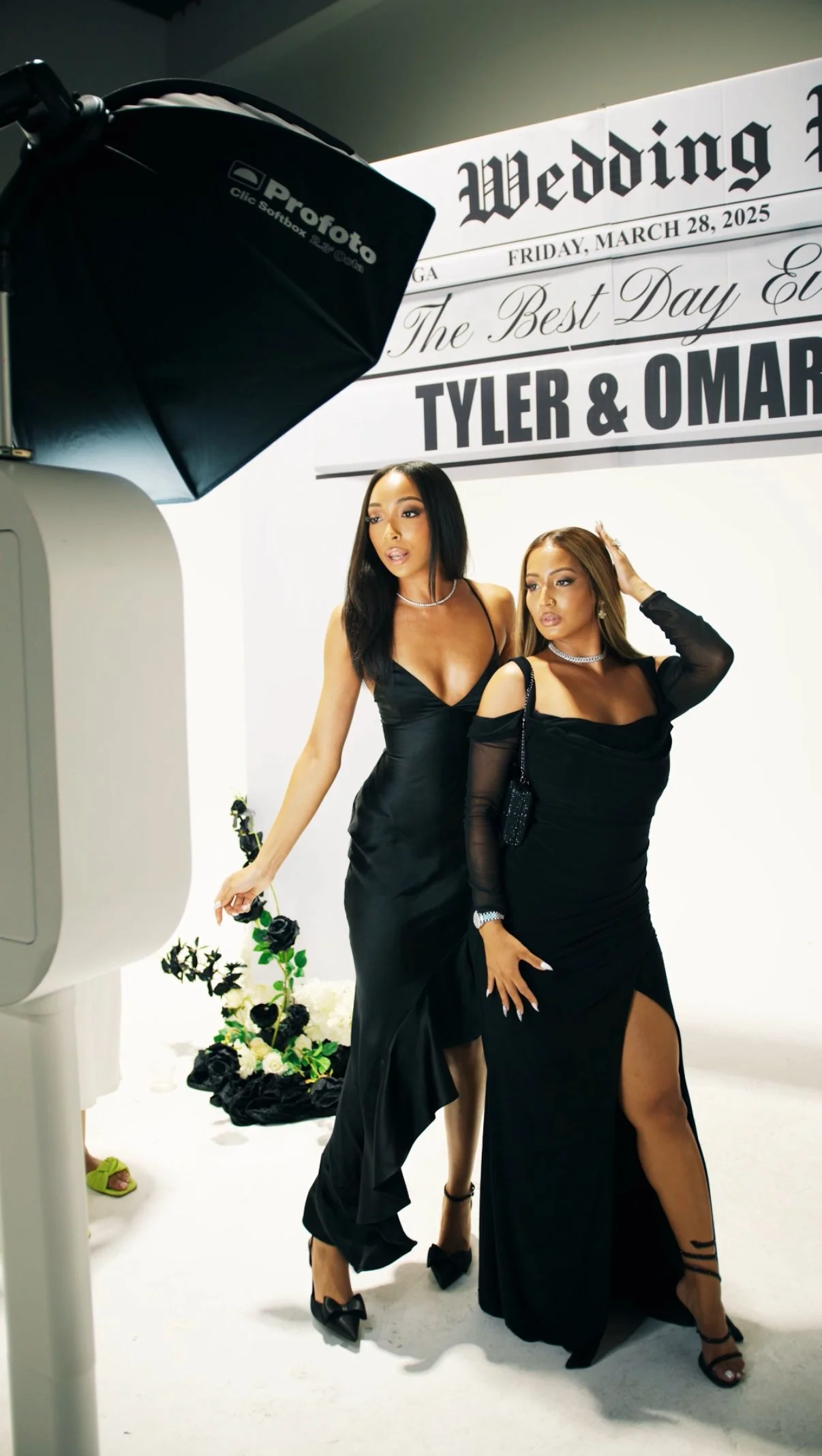 Two women in black dresses posing for a photoshoot in front of wedding-themed signage that reads "Wedding... The Best Day Ever" and the names Tyler and Omar. They are dressed elegantly, with the woman on the left holding black and white roses and the woman on the right touching her hair.