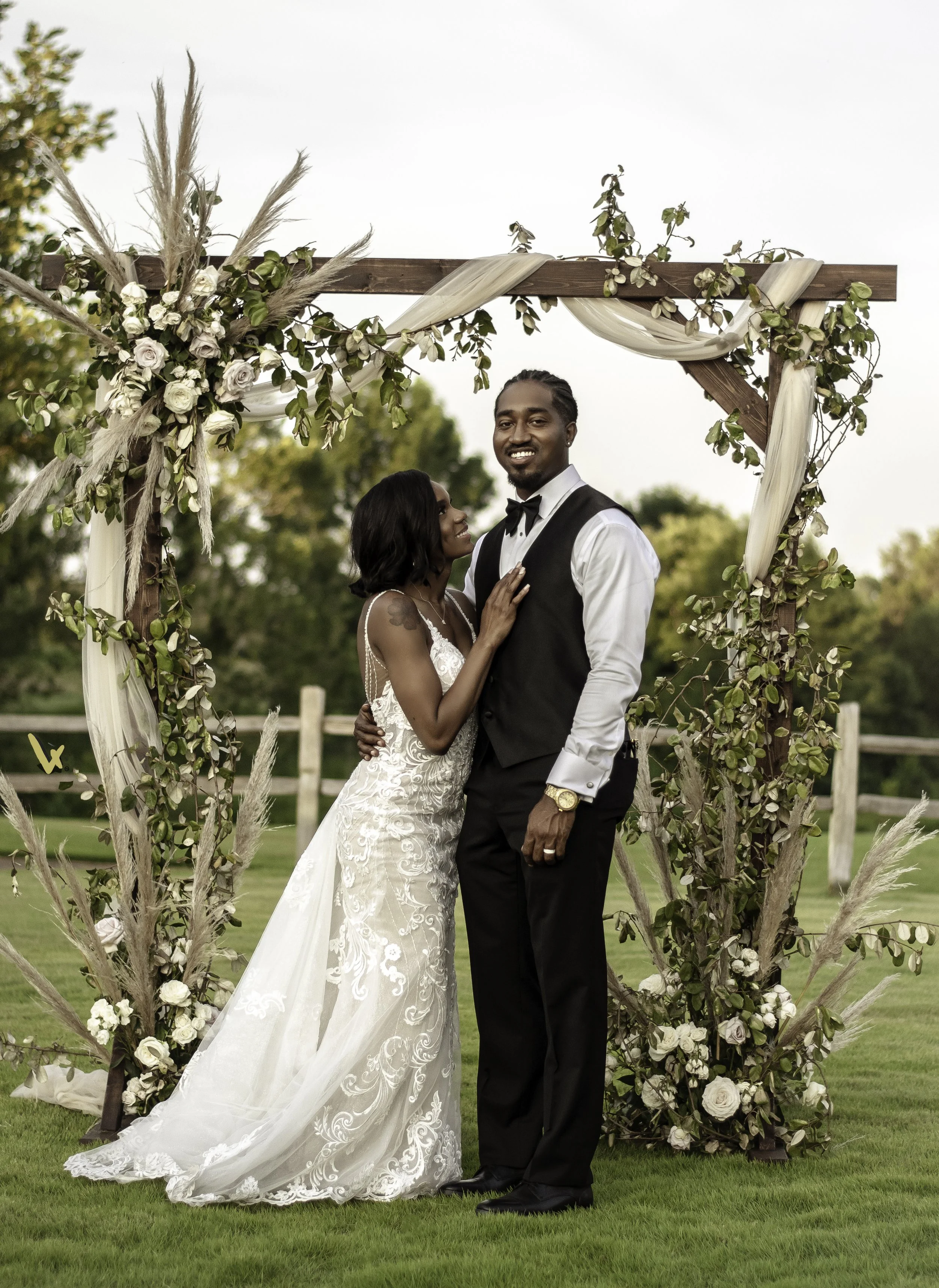 Couple in wedding attire standing under a decorated wooden arch outdoors, with greenery in the background.