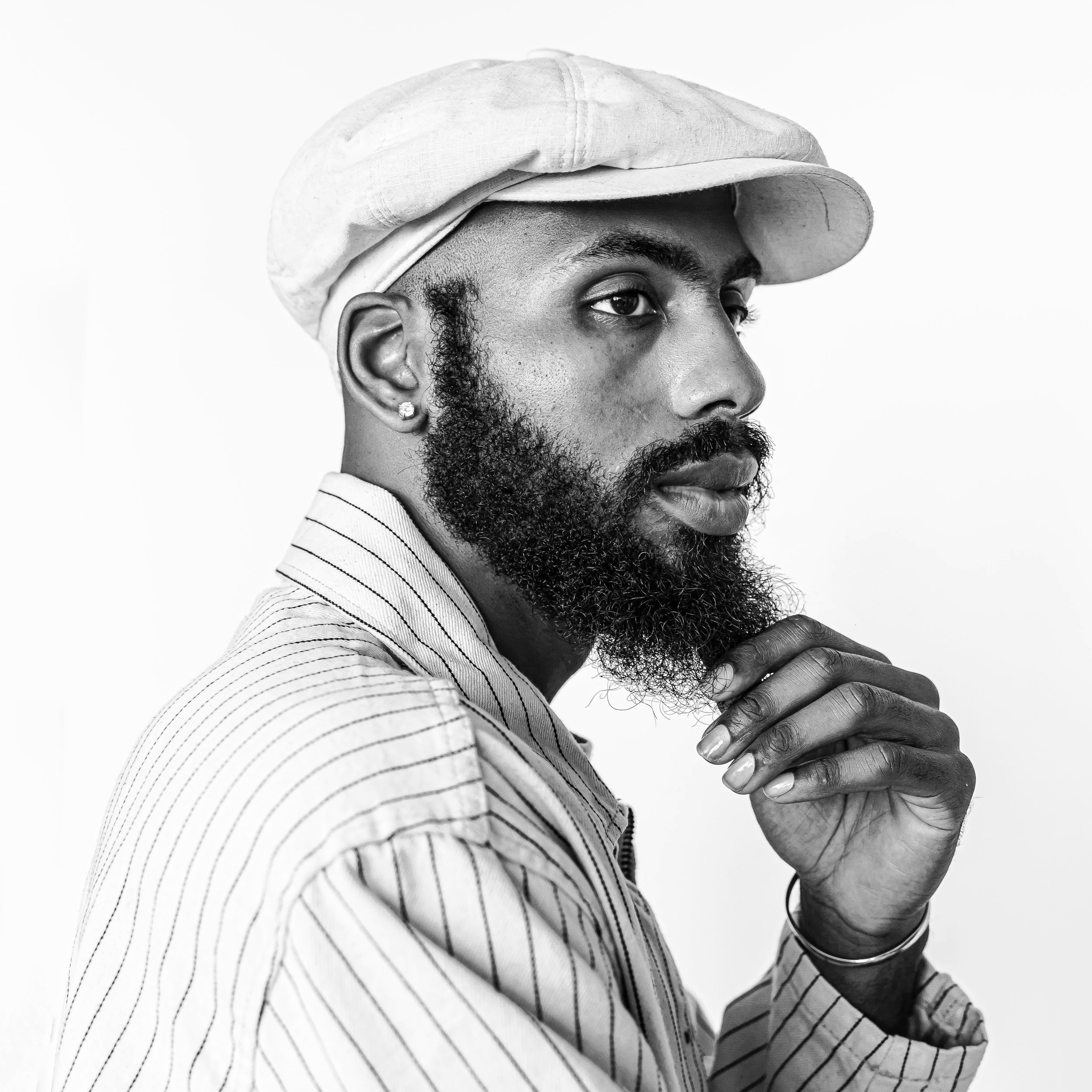 Close-up of a man with a beard and earrings, wearing a striped shirt and a cap, looking to the side, with one hand near his chest. The image is in black and white.