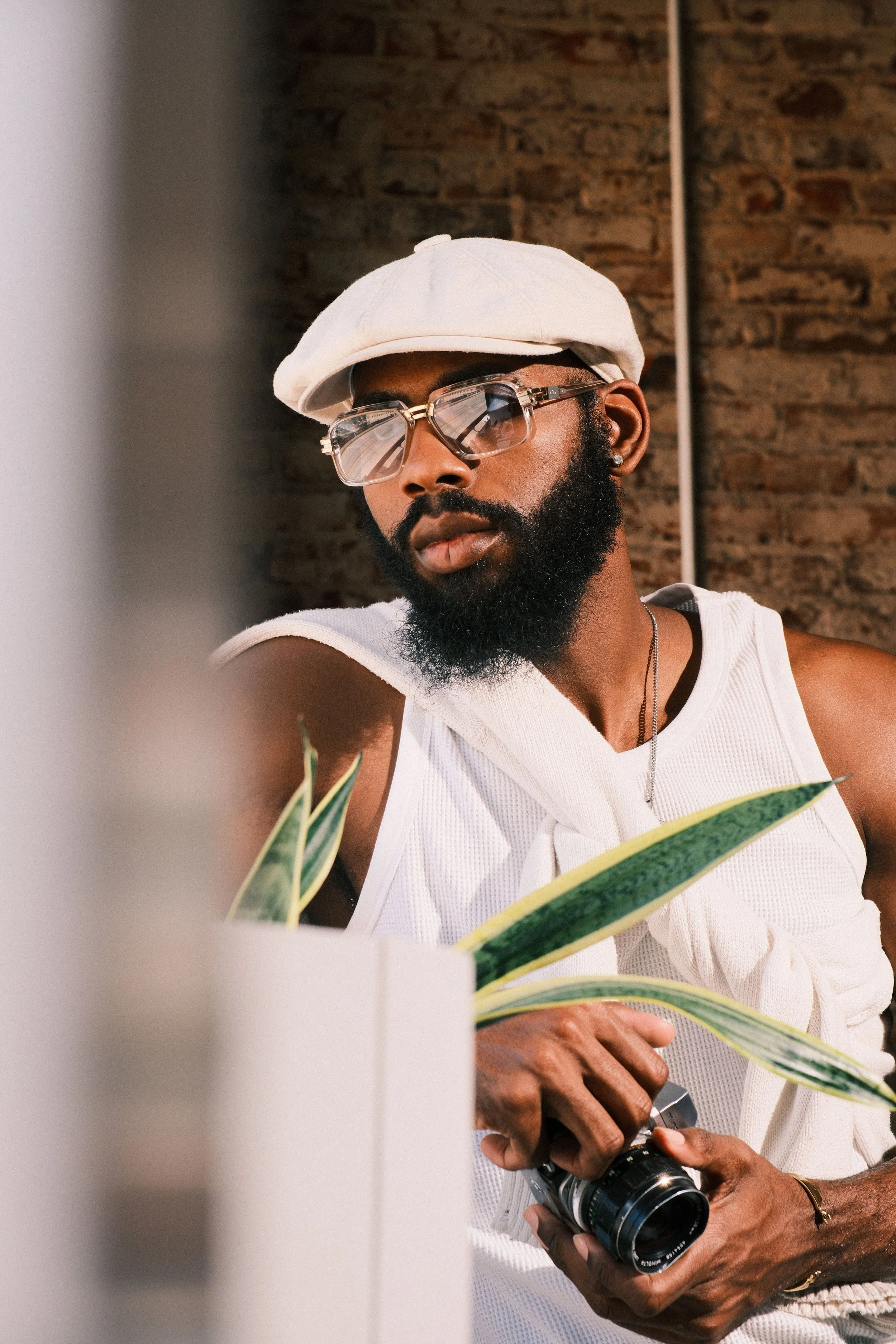 A man with dark skin, a beard, wearing sunglasses, a white hat, and a sleeveless shirt, holding a camera. The background features a brick wall.