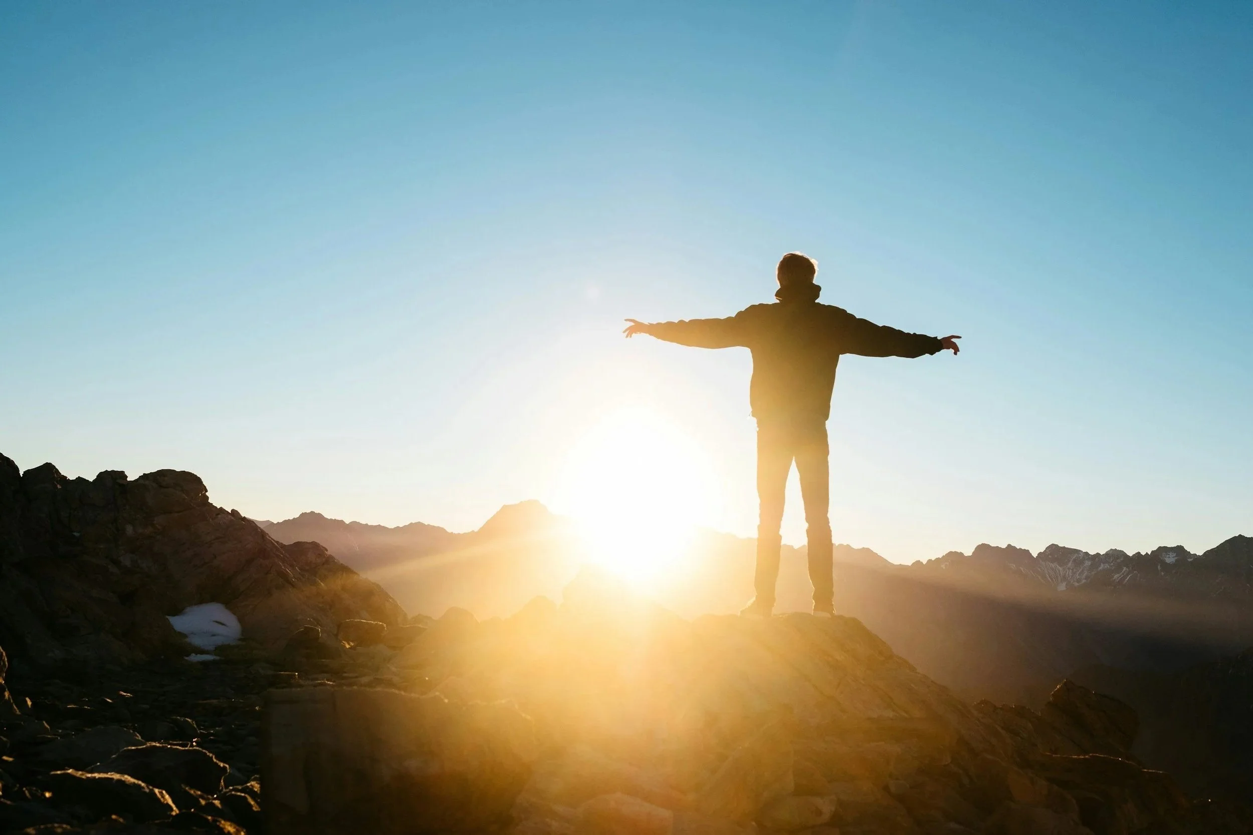 Person standing with arms outstretched on rocky terrain during sunset with mountain landscape in the background.