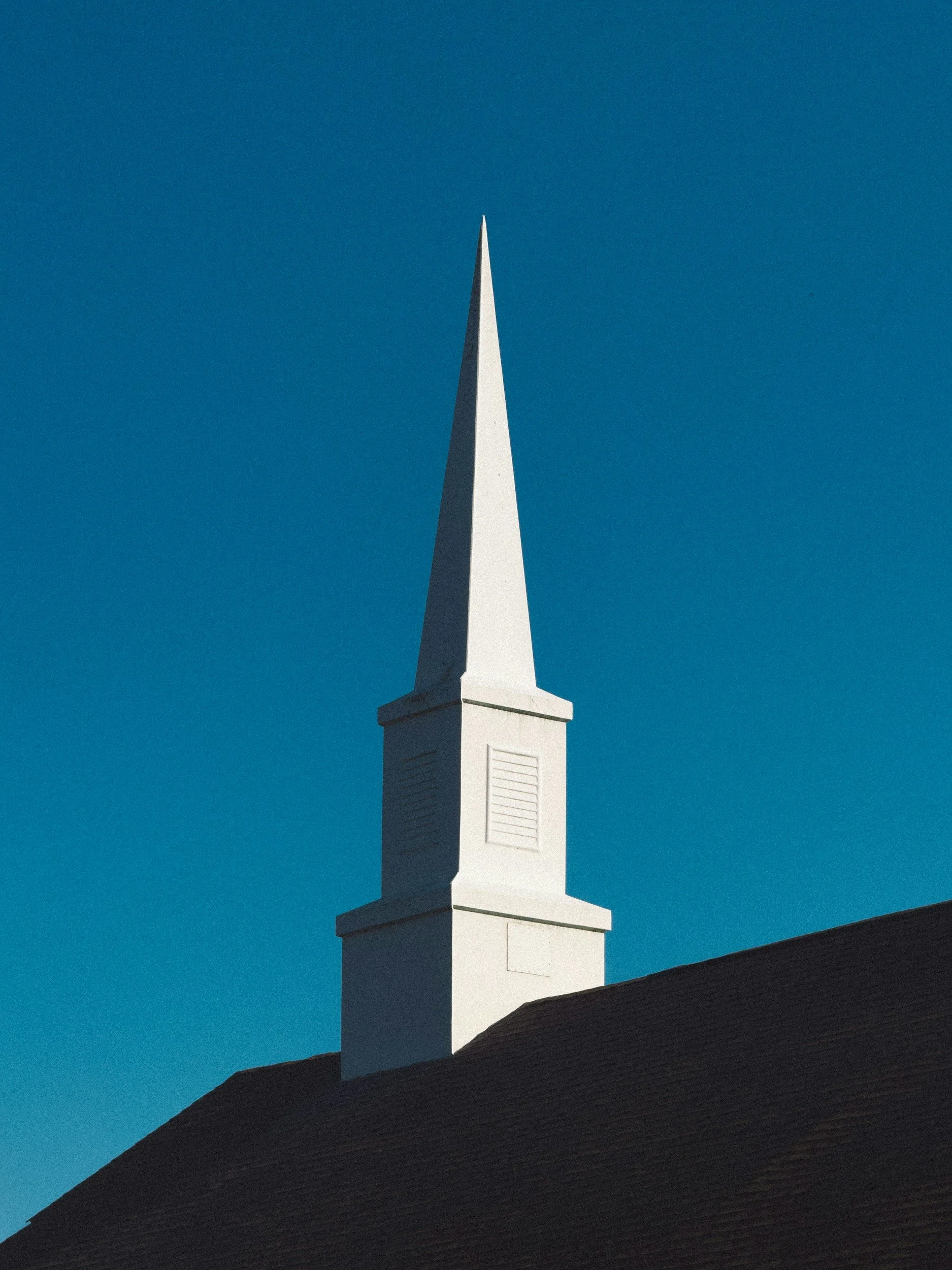 A white church steeple against a clear blue sky, with a dark roof visible at the bottom of the image.