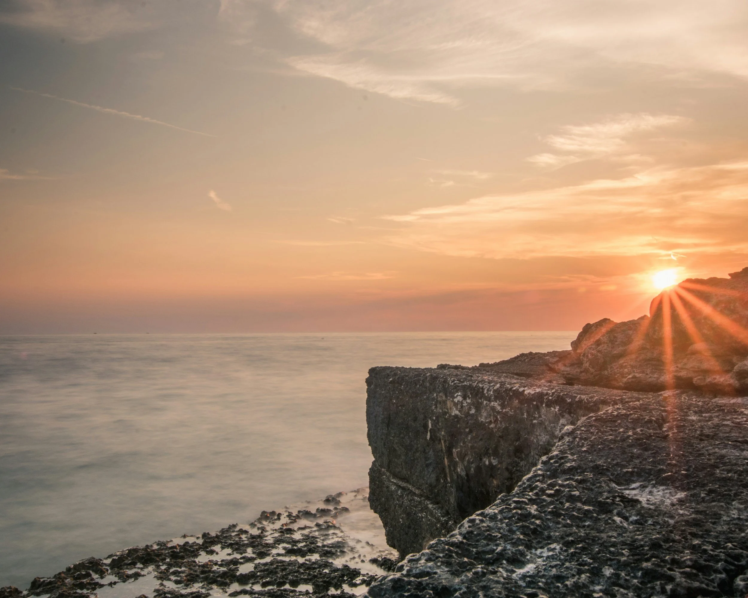 Sunset over ocean with rocky shoreline and cliff, sky with clouds and warm colors.