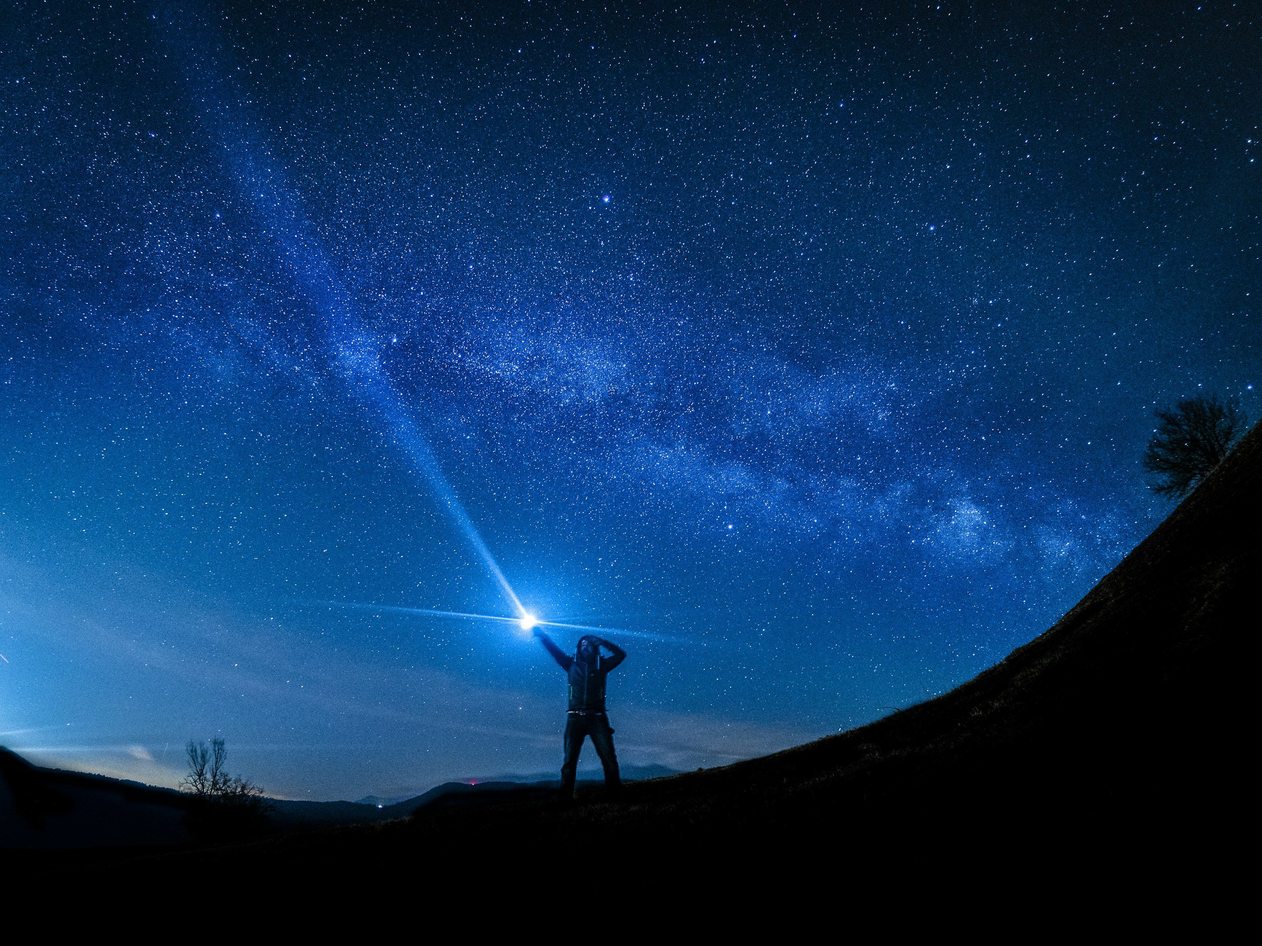 Person standing on a hill at night under a starry sky, holding a flashlight that shines upward, with trees and hills in the background.