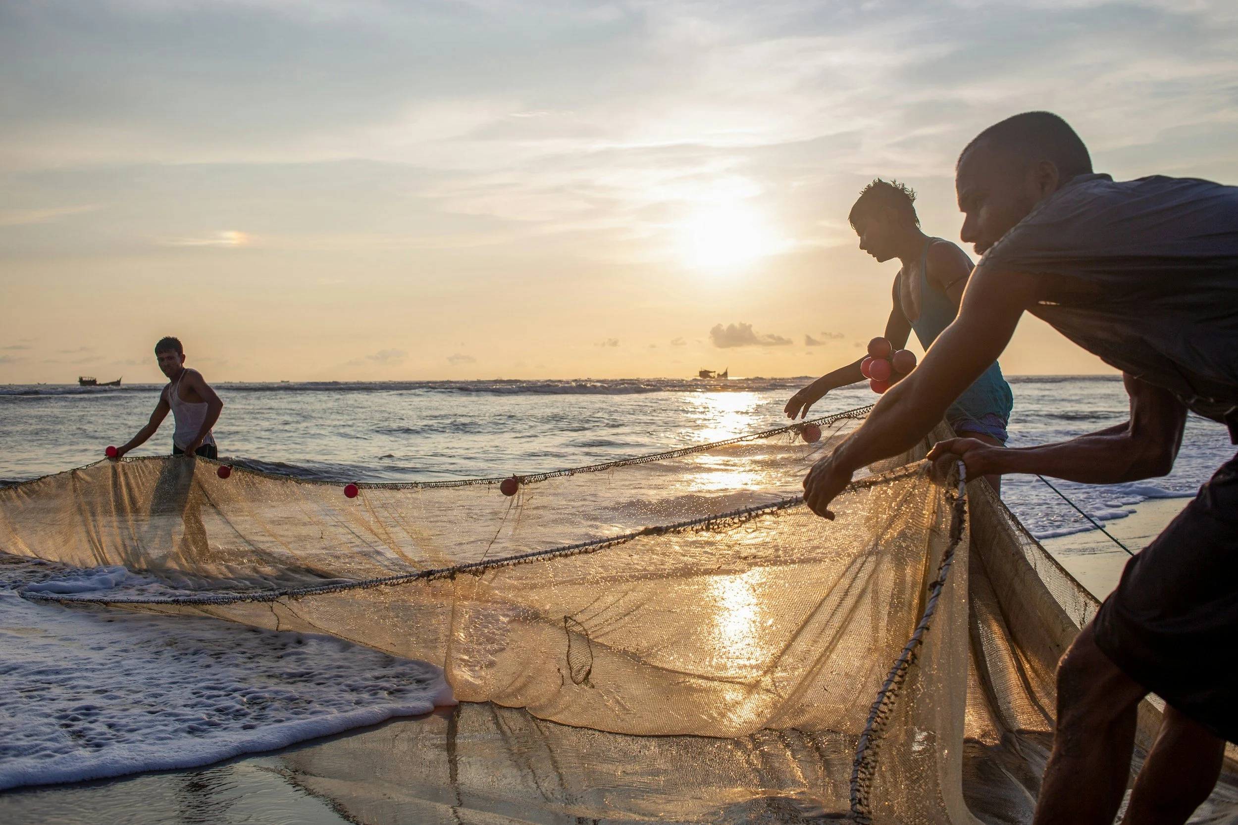Three men are fishing at the beach during sunset, pulling in a fishing net with the ocean and boats in the background.