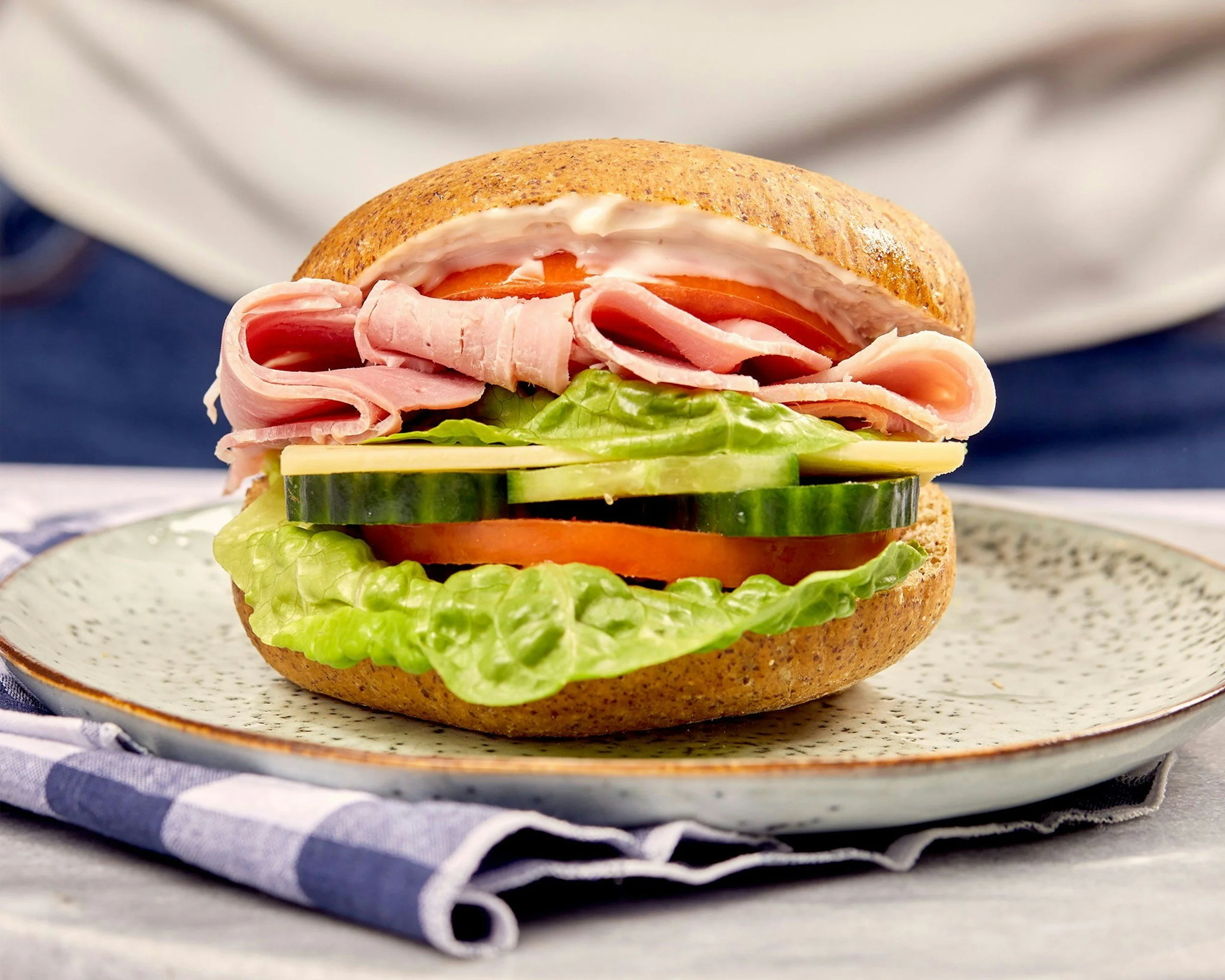 A sandwich with sliced turkey, tomato, cucumber, lettuce, and mayonnaise on a whole grain roll, served on a plate with a blue and white striped napkin.