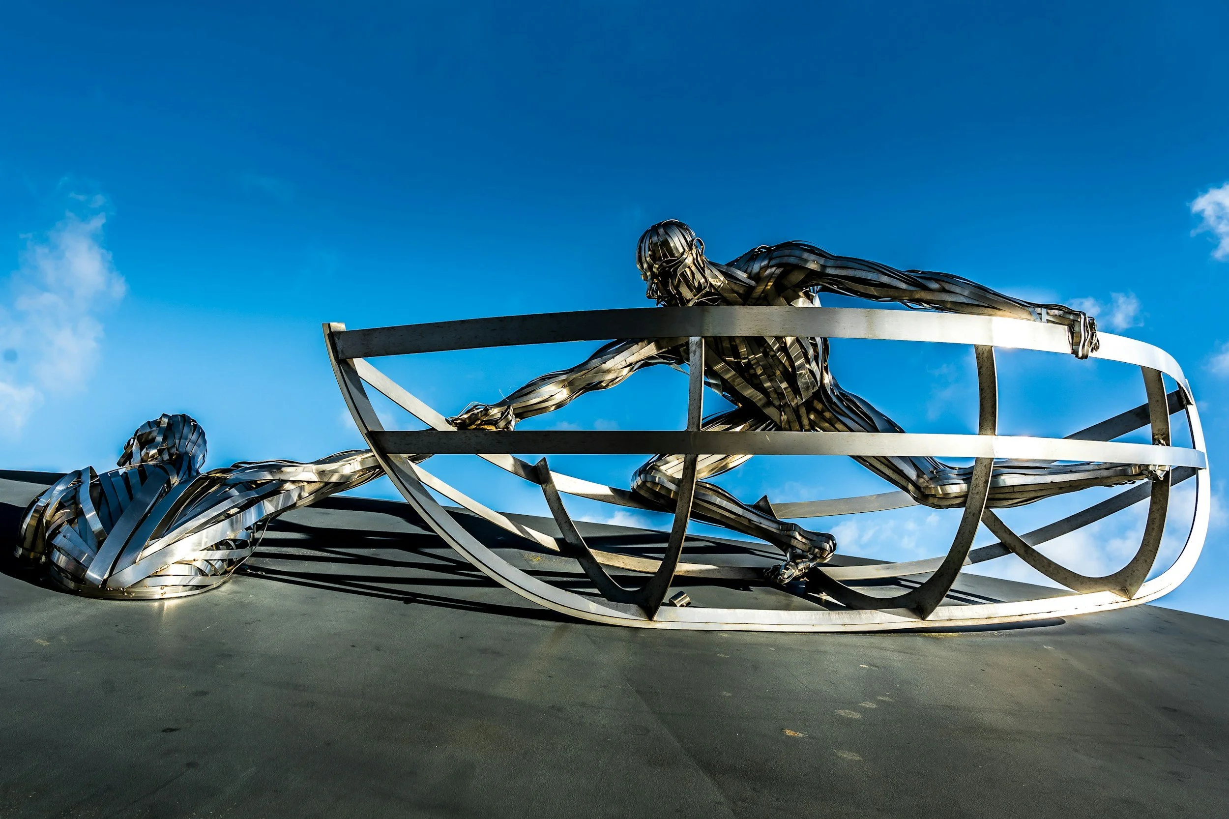 A metal sculpture of a person lying on their stomach, reaching out with one arm, with another smaller figure beside them, both part of a modern art installation against a clear blue sky.