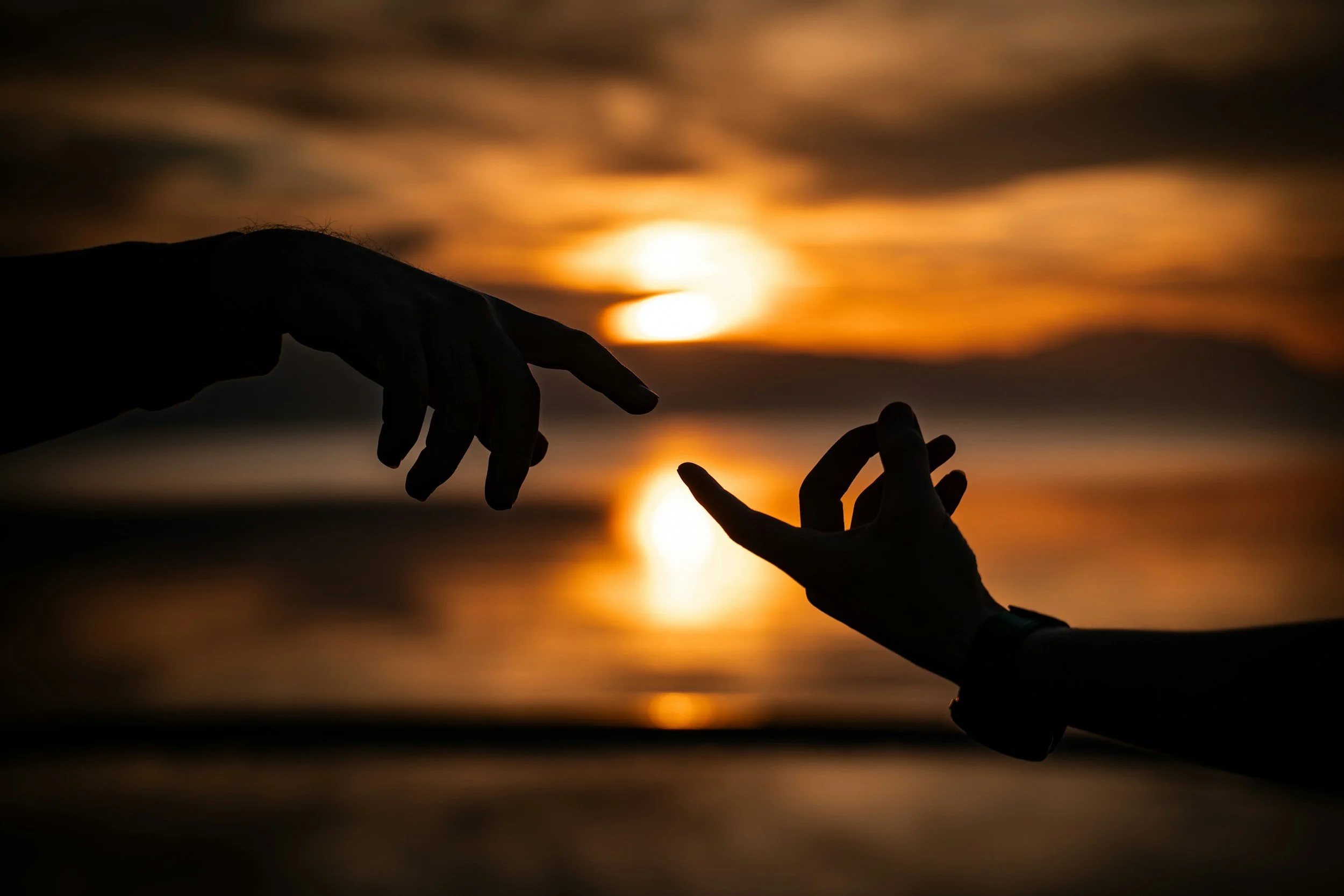 Silhouette of two hands reaching towards each other during a sunset over a body of water.