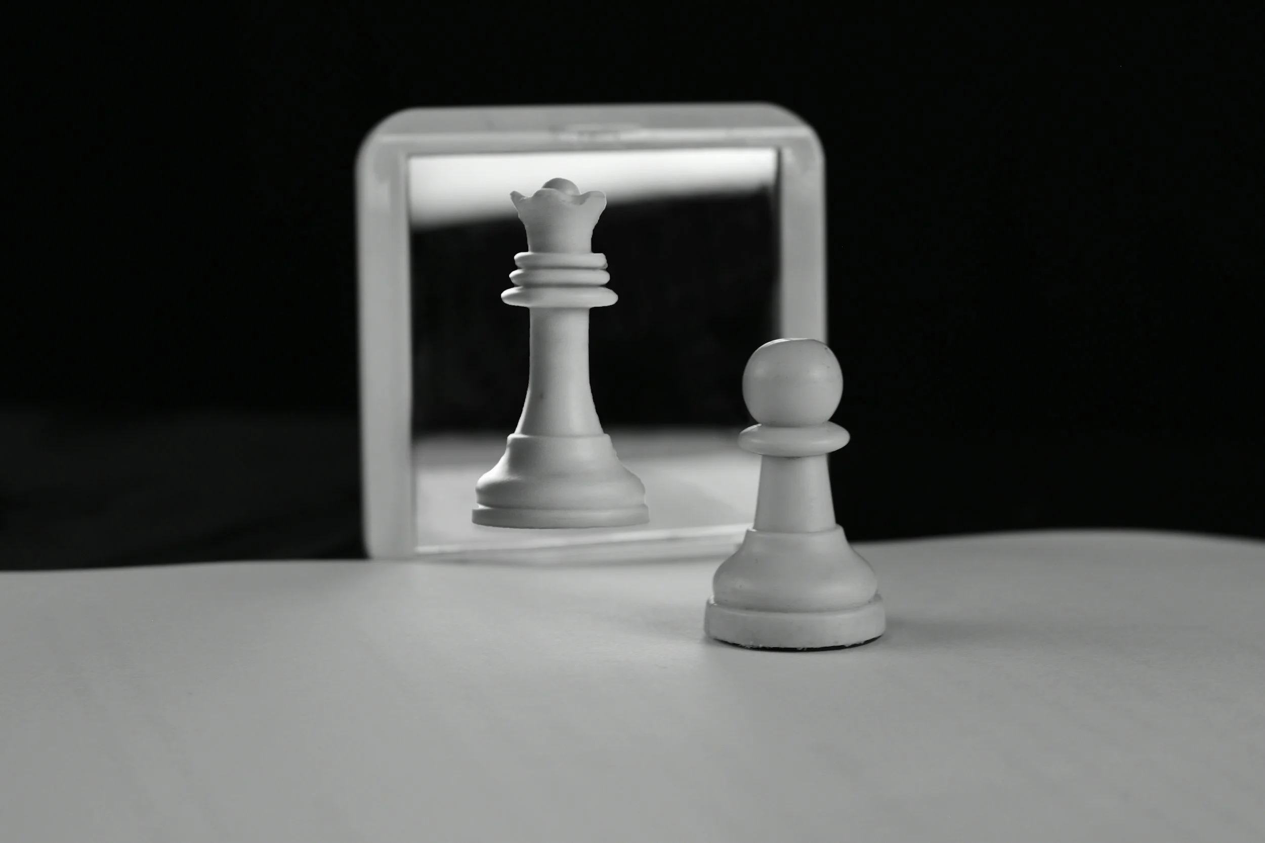 Black and white image of a chessboard with a white queen and a white pawn in front of a mirror.