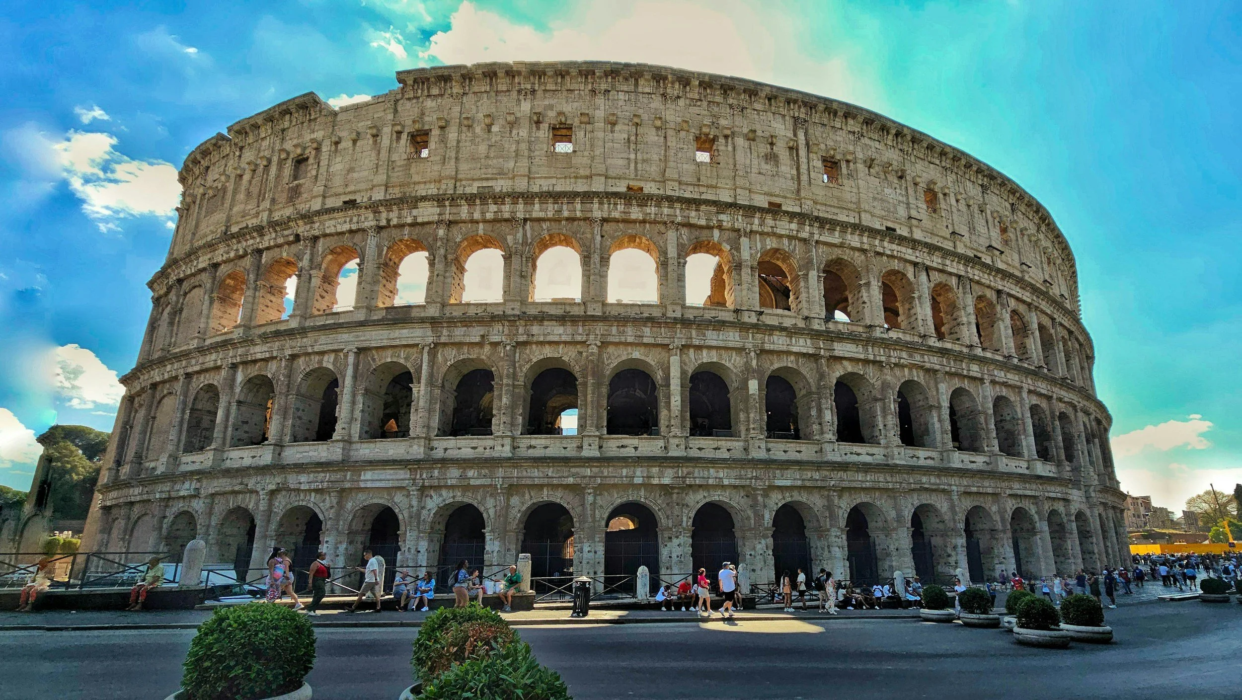 The Colosseum, an ancient Roman amphitheater, with tourists walking and sitting in front of it, under a partly cloudy sky.