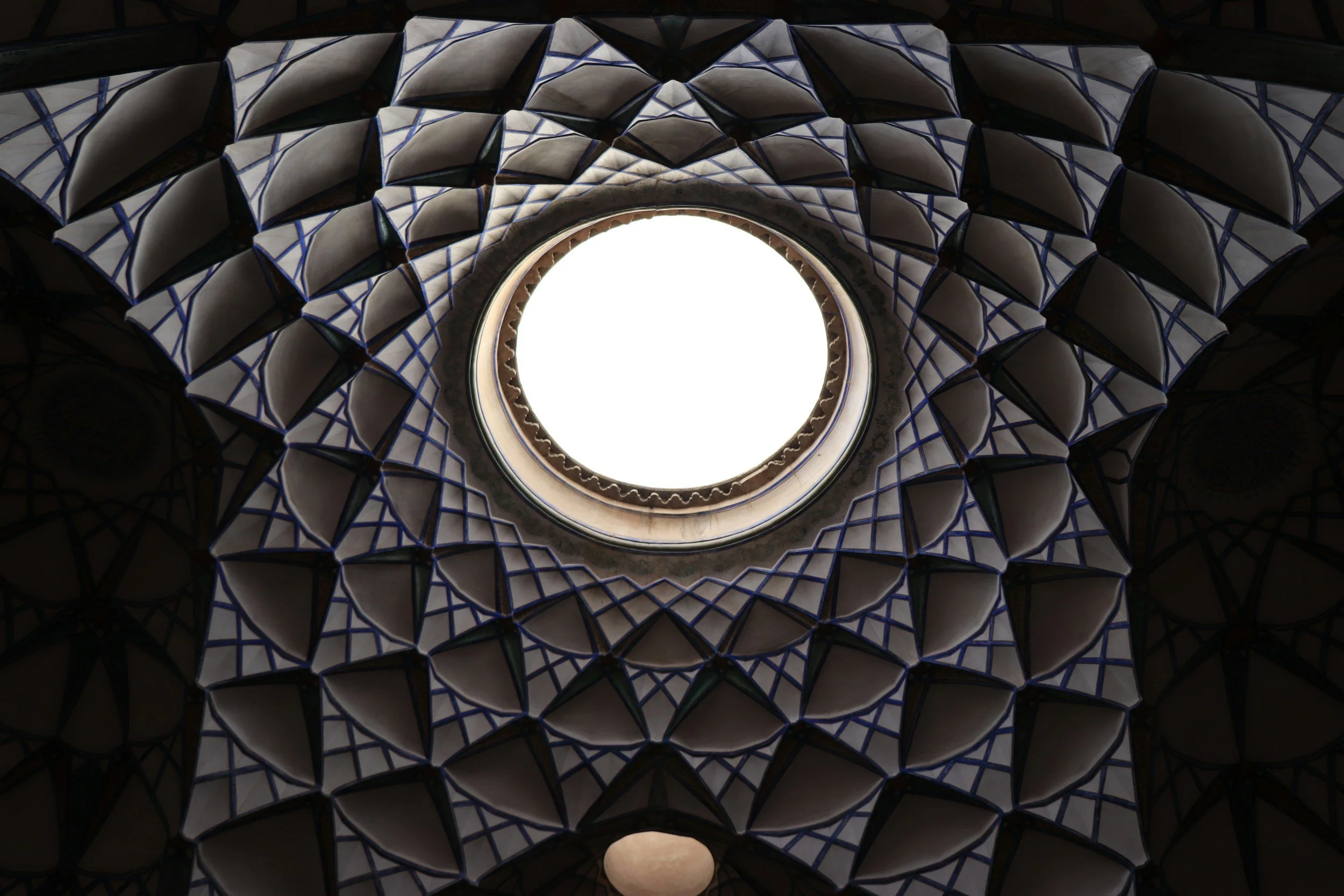 Looking up at a circular skylight with an intricate, geometric, honeycomb-like ceiling with dark framing and white filling.