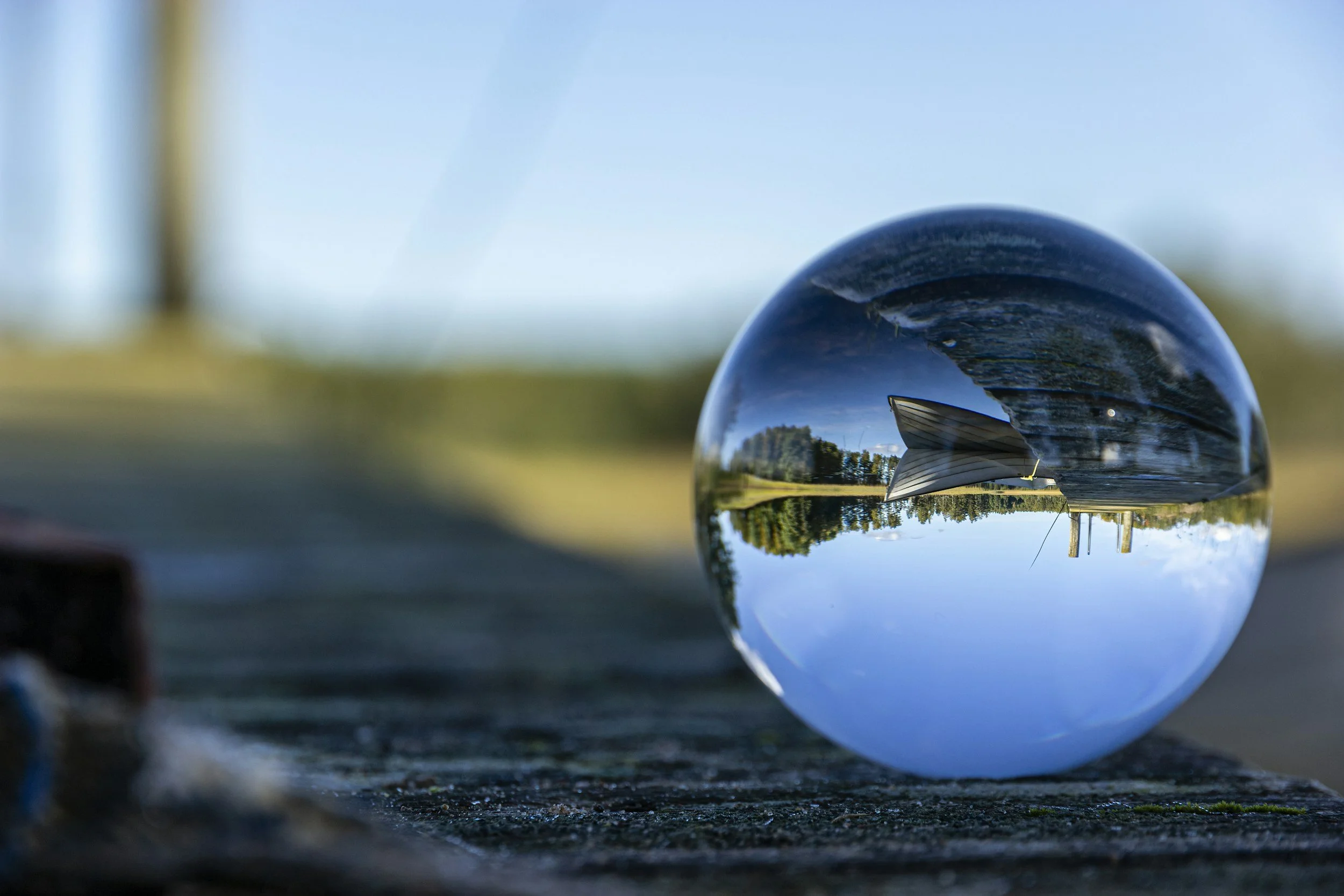 A clear glass sphere reflecting a house with a yard, trees, and a blue sky with some clouds, resting on a concrete surface outdoors.