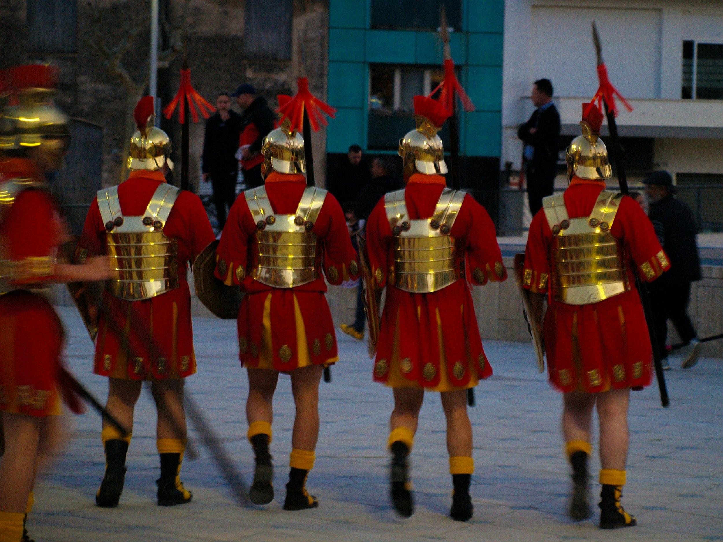 A group of people dressed in red and gold armor with helmets, walking in formation outdoors in a city street, with modern buildings and onlookers in the background.