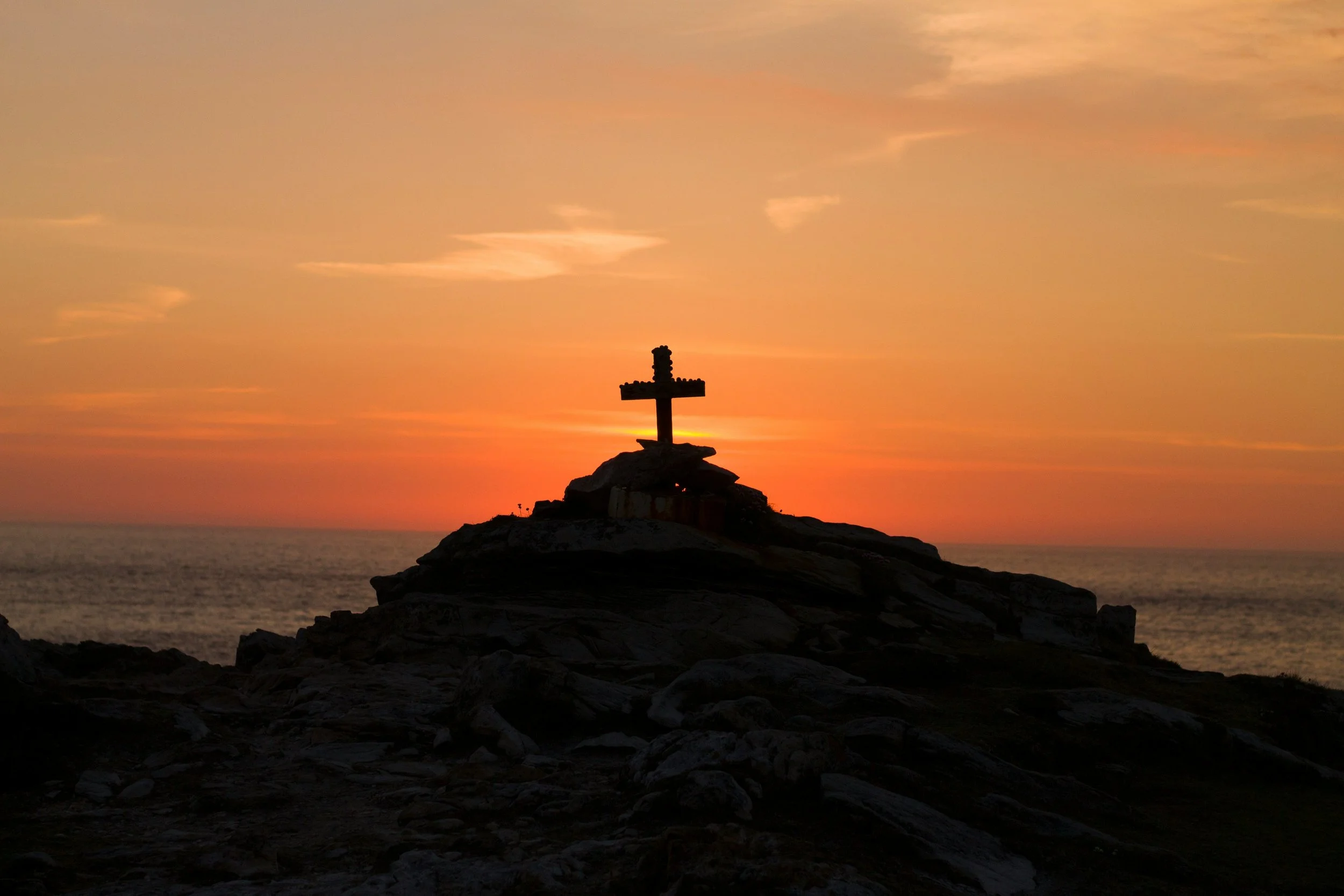 A cross sculpture on a rocky hilltop silhouetted against a colorful sunset sky over the ocean.