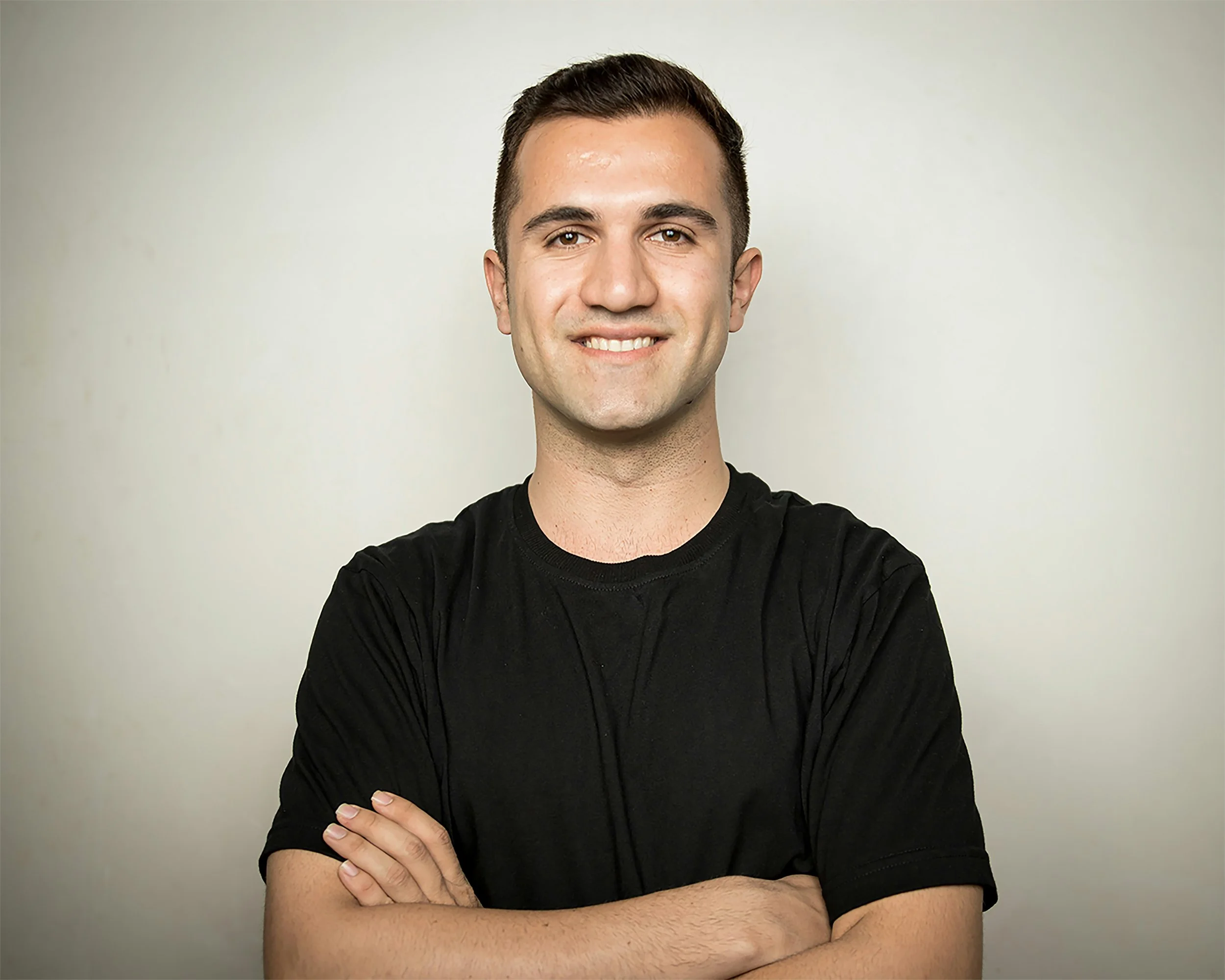 A young man with short dark hair, smiling, wearing a black shirt, crossing his arms, standing against a plain off-white background.