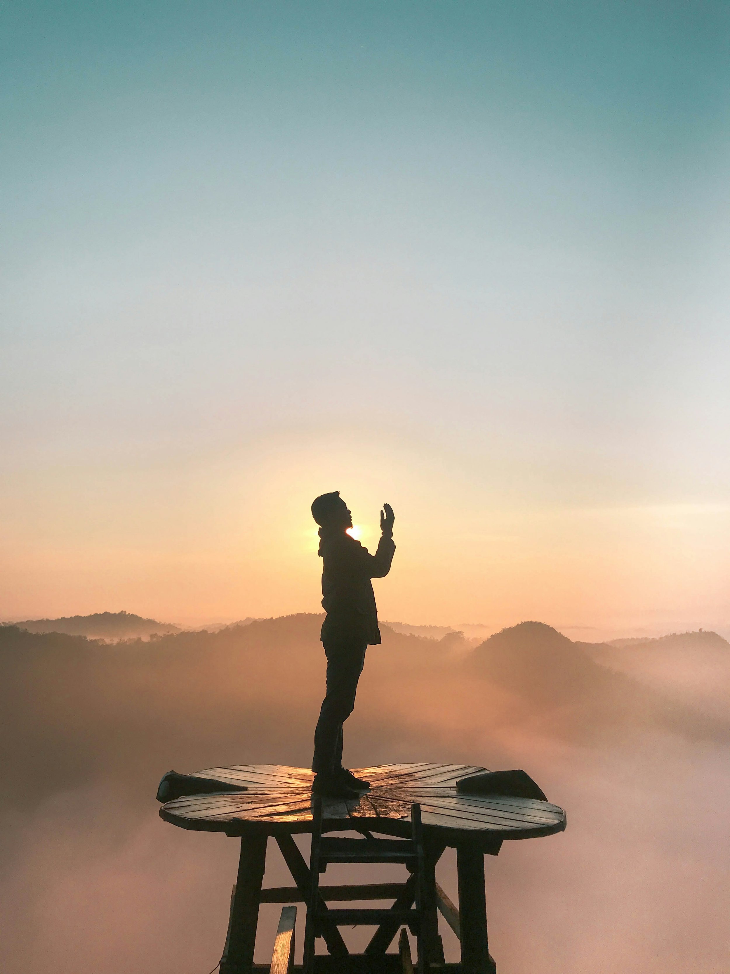 A person standing on a circular wooden platform during sunset, with mountains and a misty landscape in the background.