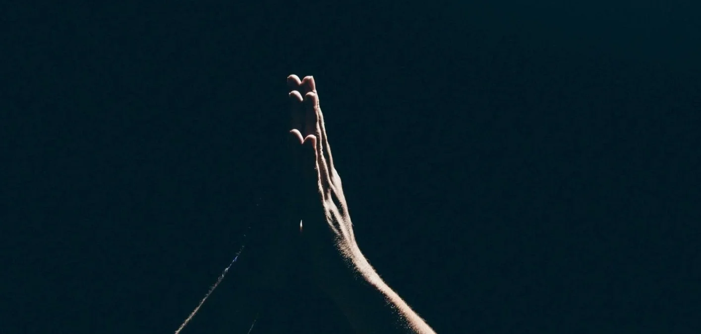 Close-up of a person's hand pressed together in prayer or greeting, with a dark background.