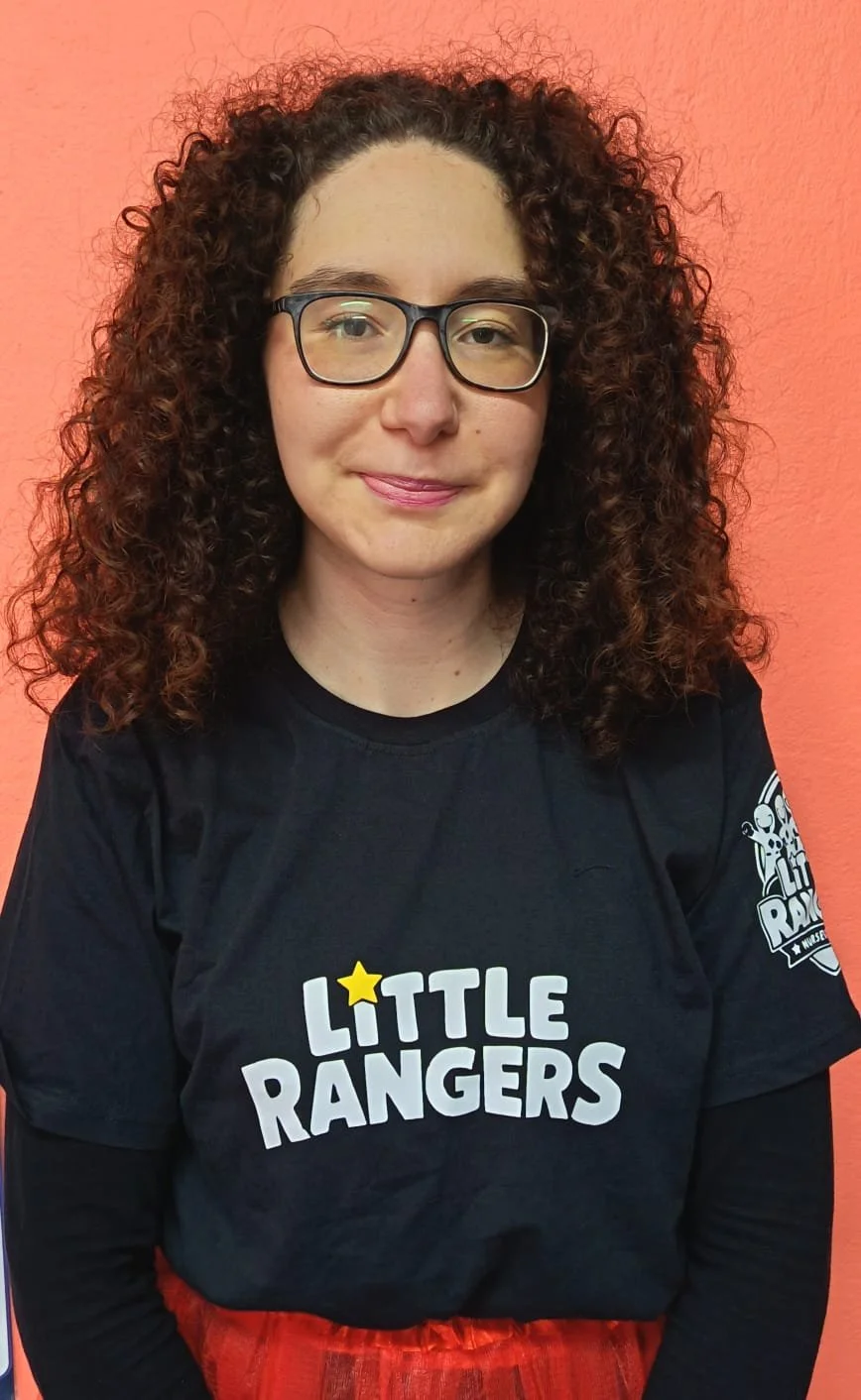 A woman with curly brown hair, glasses, and light skin, smiling while facing the camera, wearing a black 'Little Rangers' T-shirt against a peach-colored wall.
