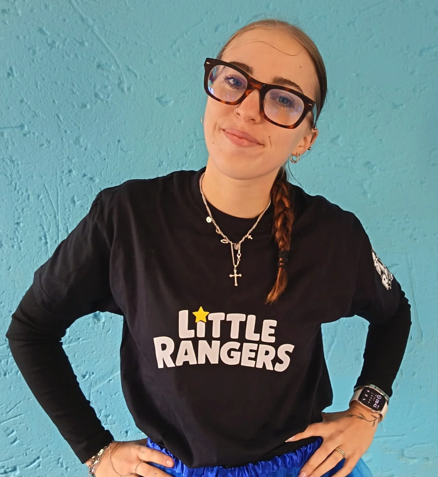 Young woman with glasses and braided hair standing against a turquoise wall, wearing a black long-sleeve shirt with "Little Rangers" printed on it, multiple necklaces, a smartwatch, and silver earrings.