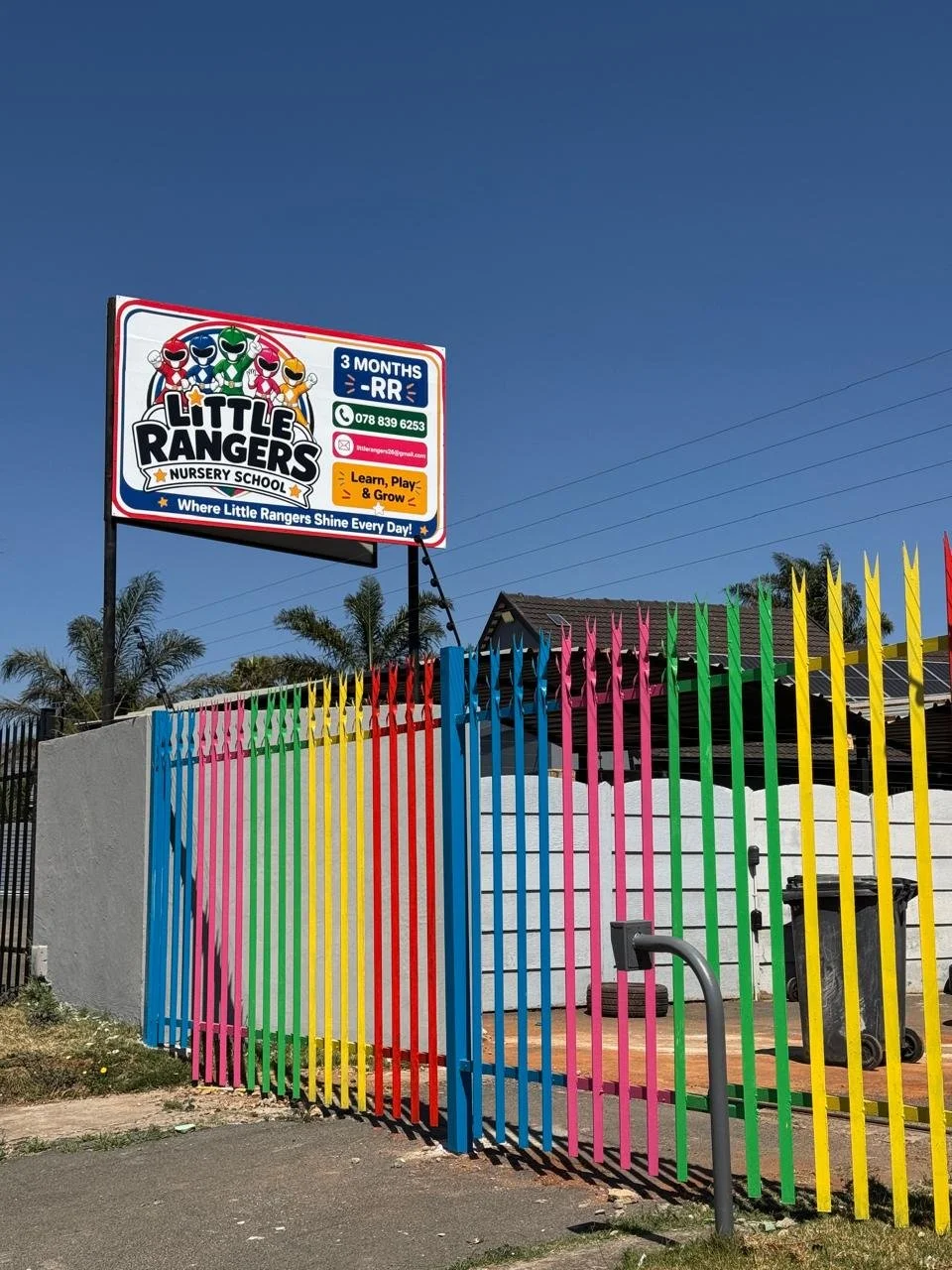 Colorful metal gate in front of a nursery school with a sign displaying the school's name 'Little Rangers Nursery School' and contact information, under a clear blue sky.