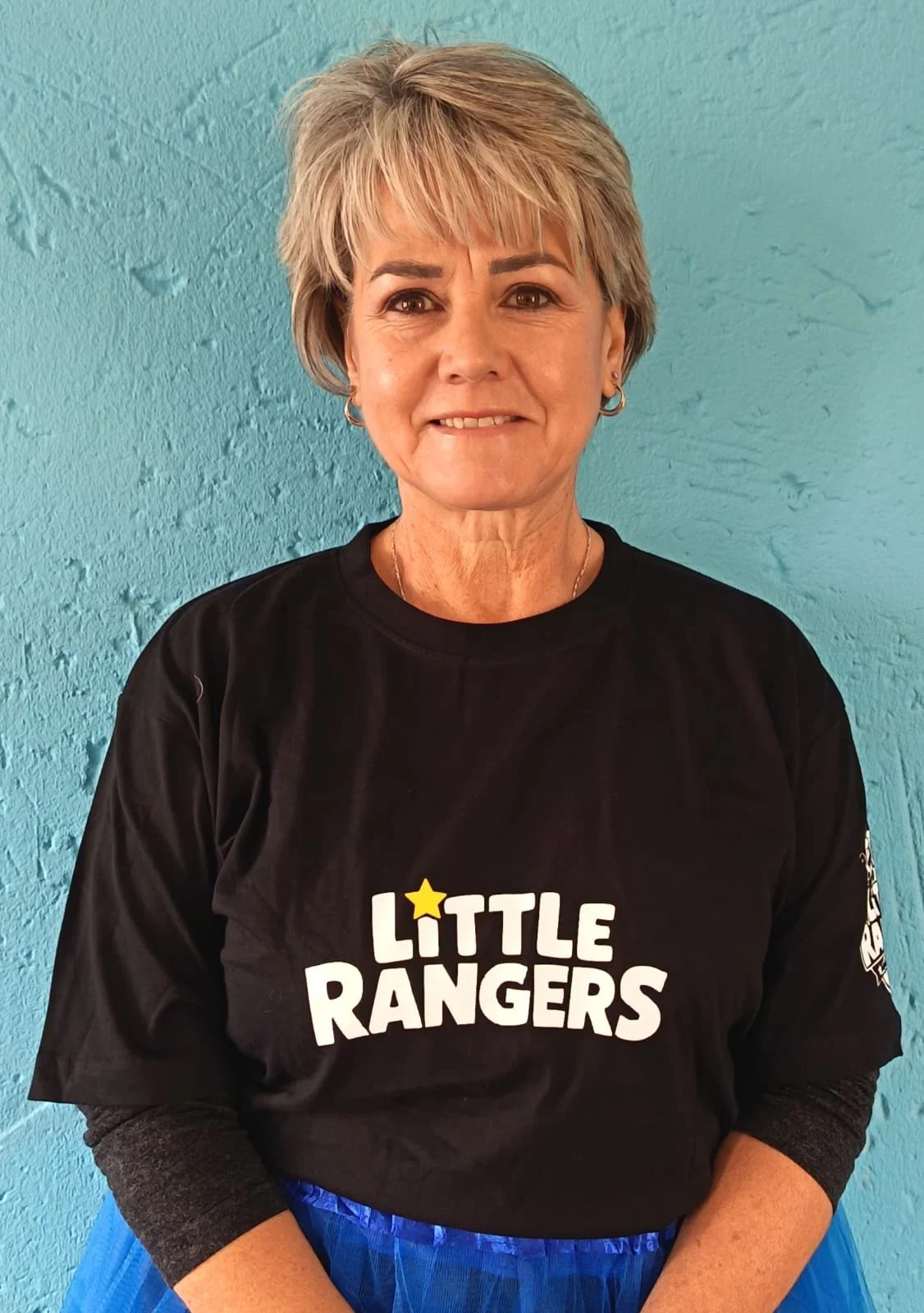 A woman with short, gray hair smiling at the camera, wearing a black t-shirt with the words 'LITTLE RANGERS' printed on it, standing against a light blue textured wall.
