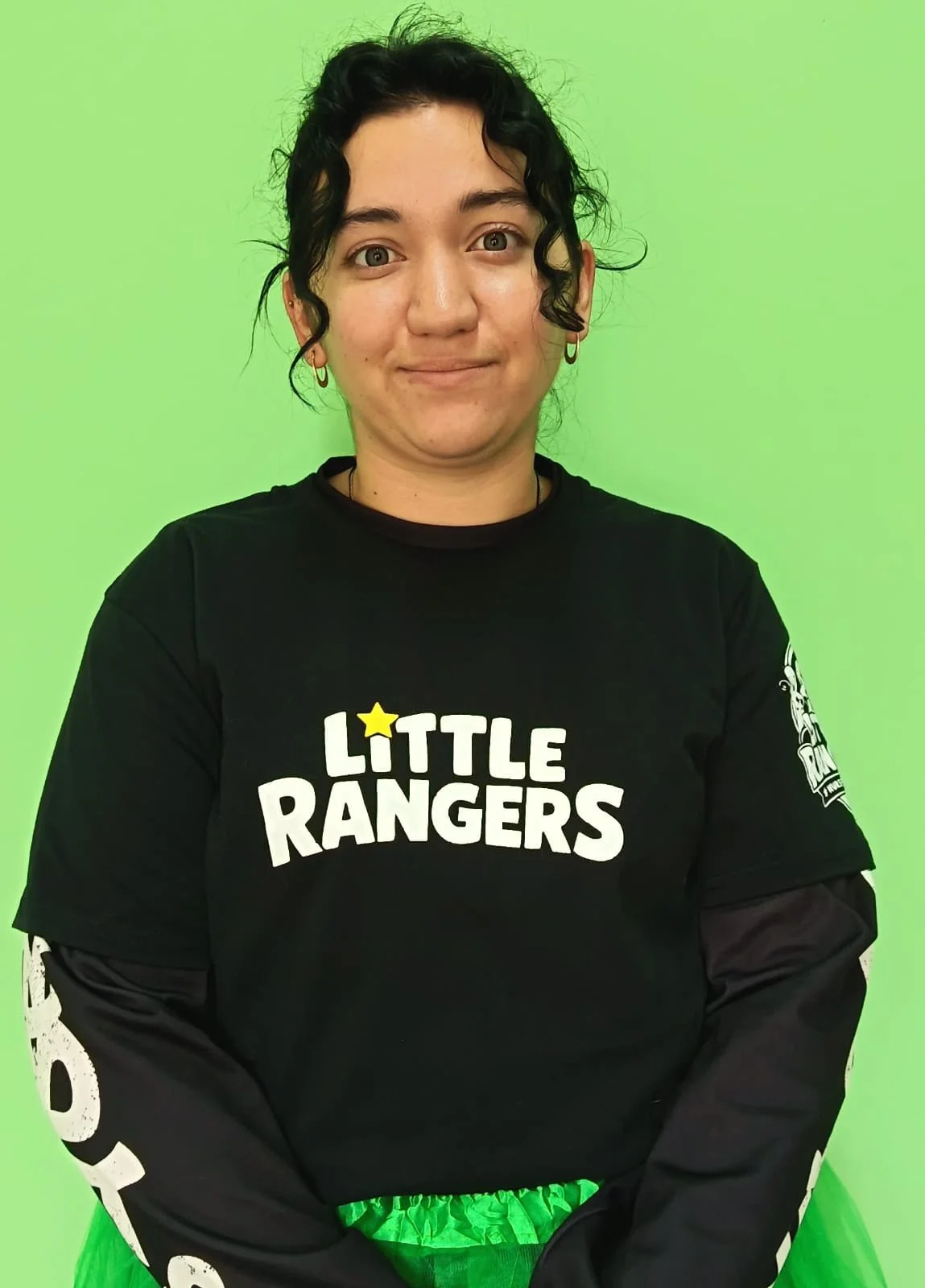 A person with short curly black hair and hoop earrings stands against a lime green background, wearing a black shirt with 'Little Rangers' text and a yellow star, along with a black long-sleeve underneath.