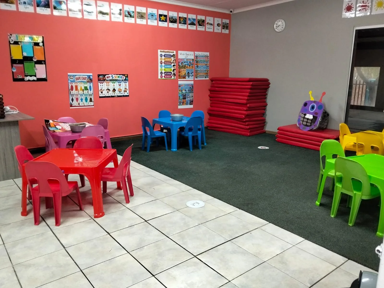Colorful children's playroom with small tables and chairs, decorated walls with educational posters, and toys including a purple monster face, stacked red mats, and a window with a railing.