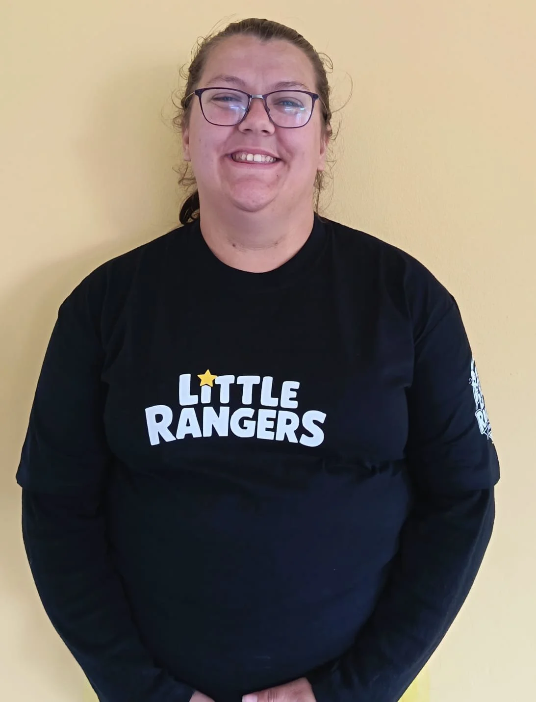 A woman with glasses and long hair smiling, wearing a black shirt with the text 'Little Rangers' and a star logo, standing against a yellow wall.