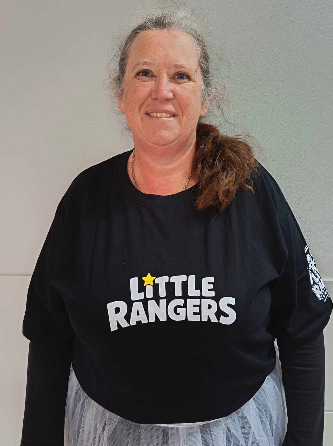 A woman with gray hair in a ponytail, wearing a black 'Little Rangers' t-shirt, smiles while standing against a plain wall.