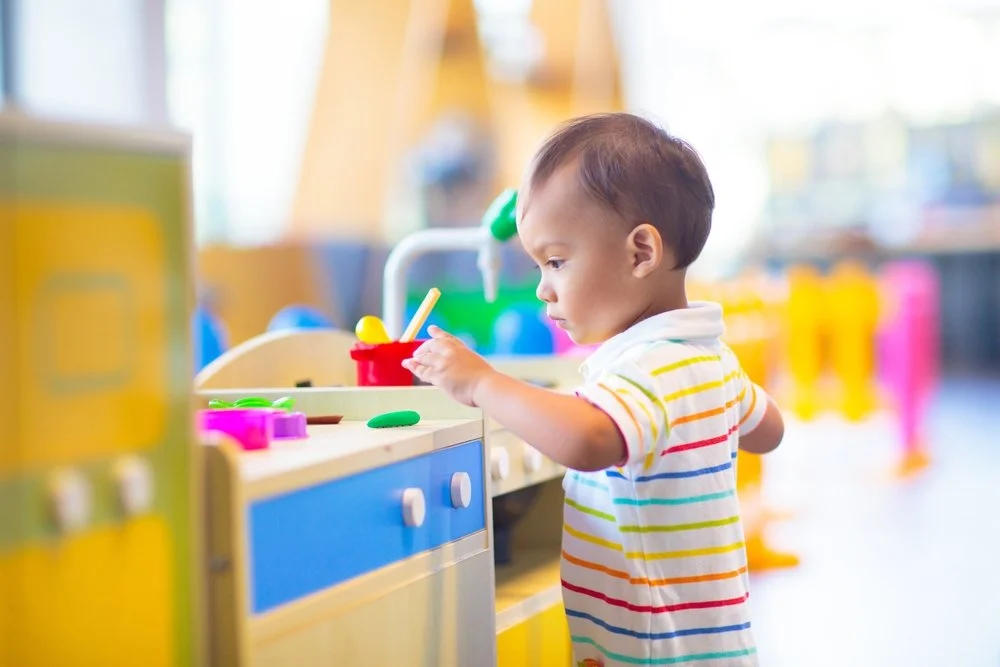 A young child playing with colorful toy kitchen utensils in a play kitchen set in a brightly lit room.