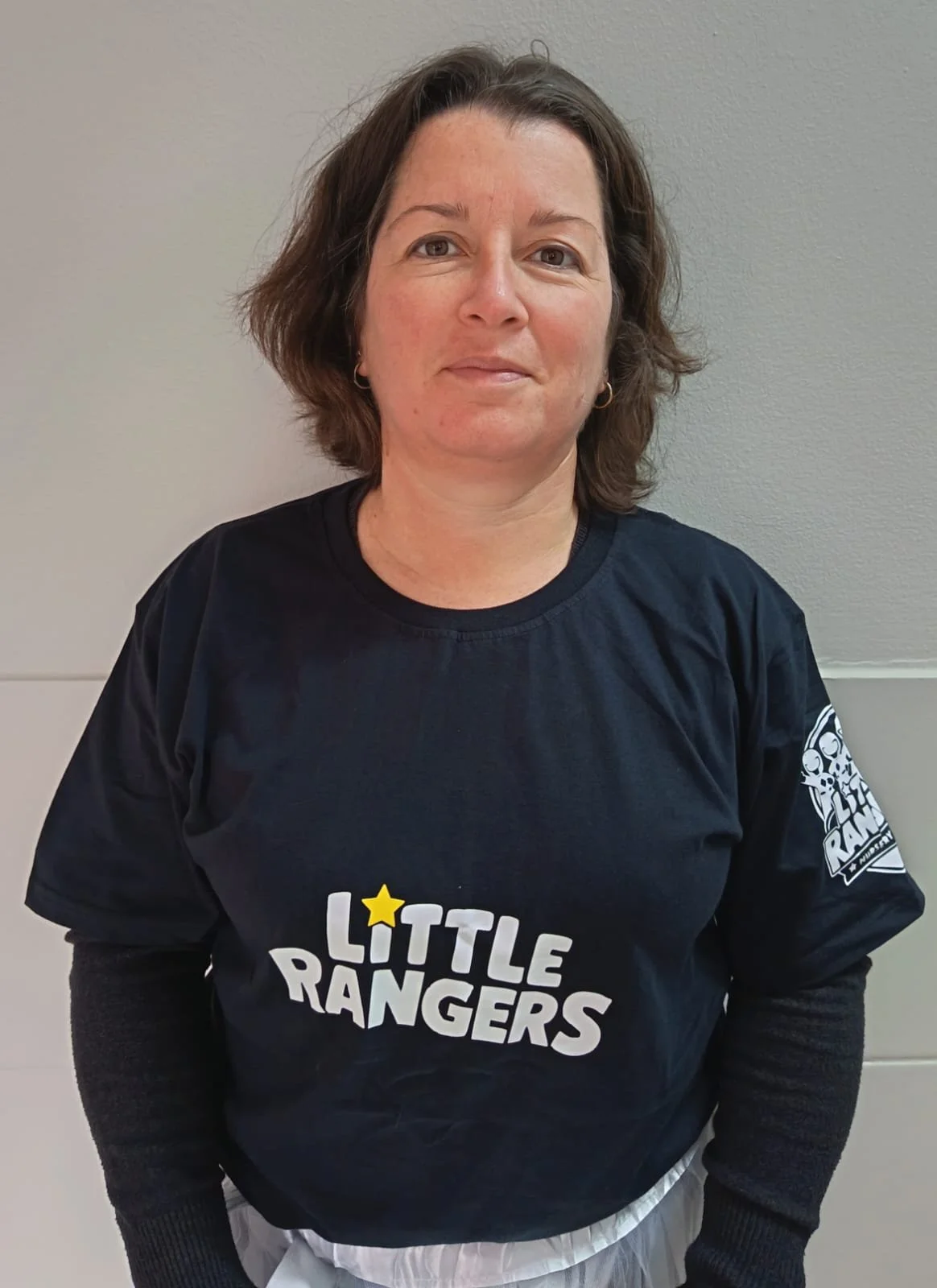 Woman with short brown hair wearing a black 'Little Rangers' T-shirt and striped pants, standing against a light gray wall.
