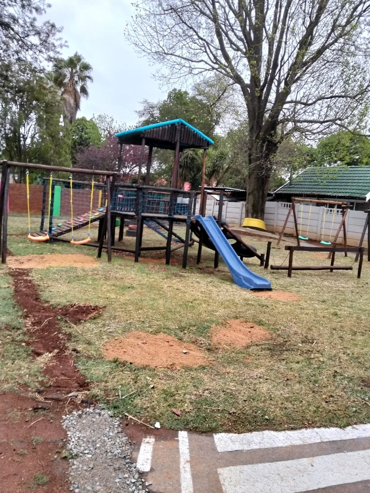 Children's playground with swings, a slide, a sandbox, and a climbing structure in a grassy backyard with trees and a white fence.