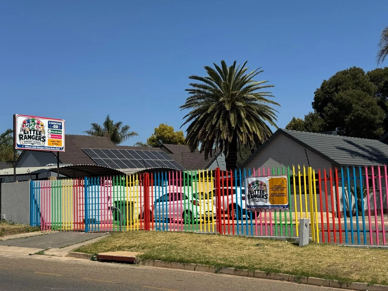 Colorful fence surrounding Little Rangers Nursery School, with cars parked behind, palm trees, and solar panels on a house roof, under a clear blue sky.