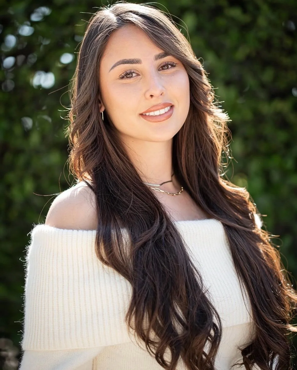 A young woman with long, wavy brown hair and light skin, smiling outdoors with green foliage in the background. She is wearing an off-the-shoulder cream sweater, hoop earrings, layered necklaces, and has natural makeup.