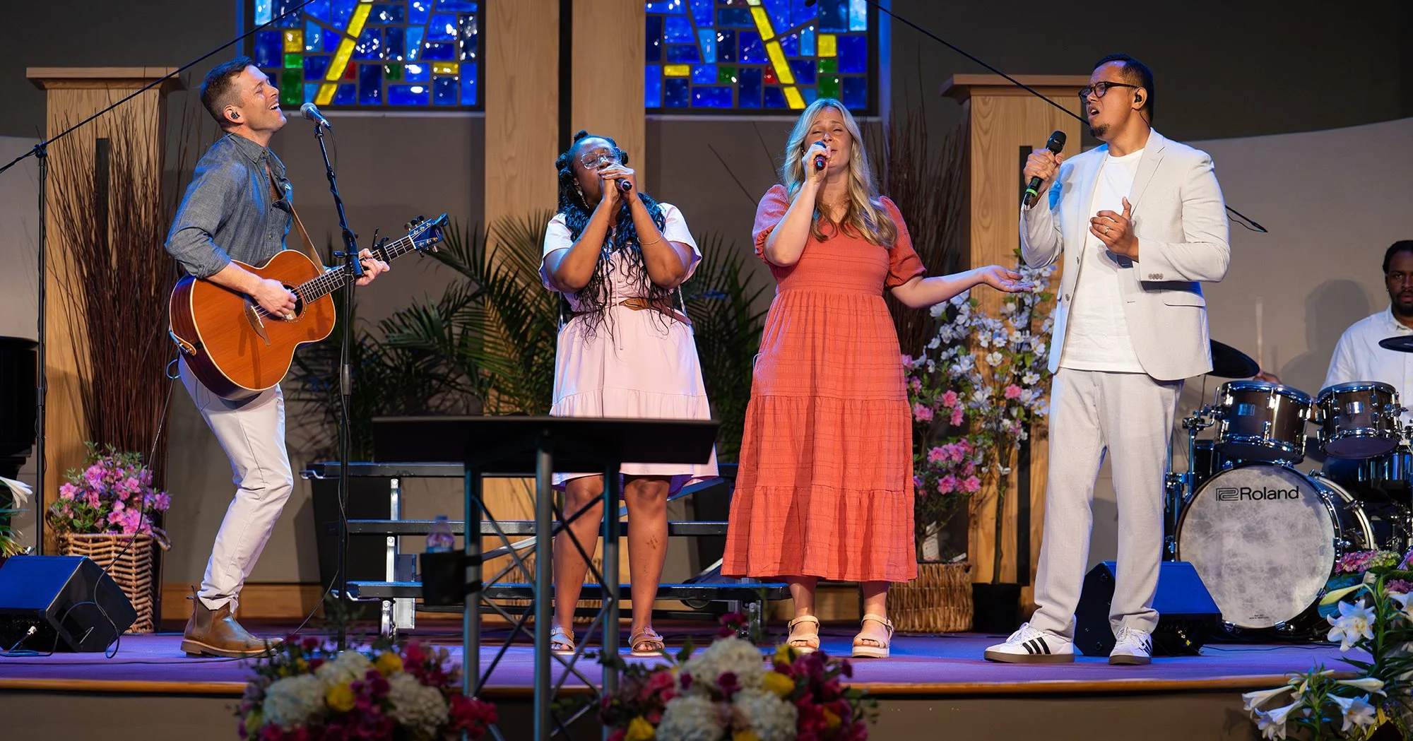 A group of five people singing and playing musical instruments on a stage decorated with flowers and greenery, with a stained glass window background.