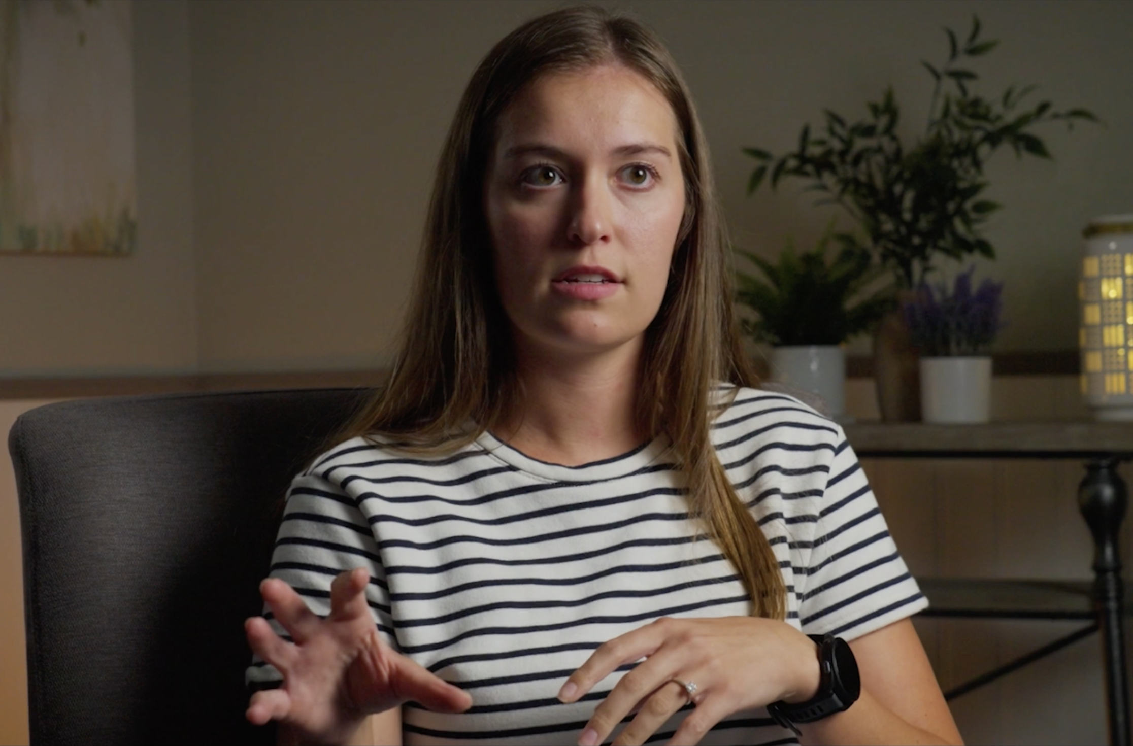 A woman with long brown hair wearing a black and white striped shirt, talking with her hands in a room with potted plants and a wall hanging.
