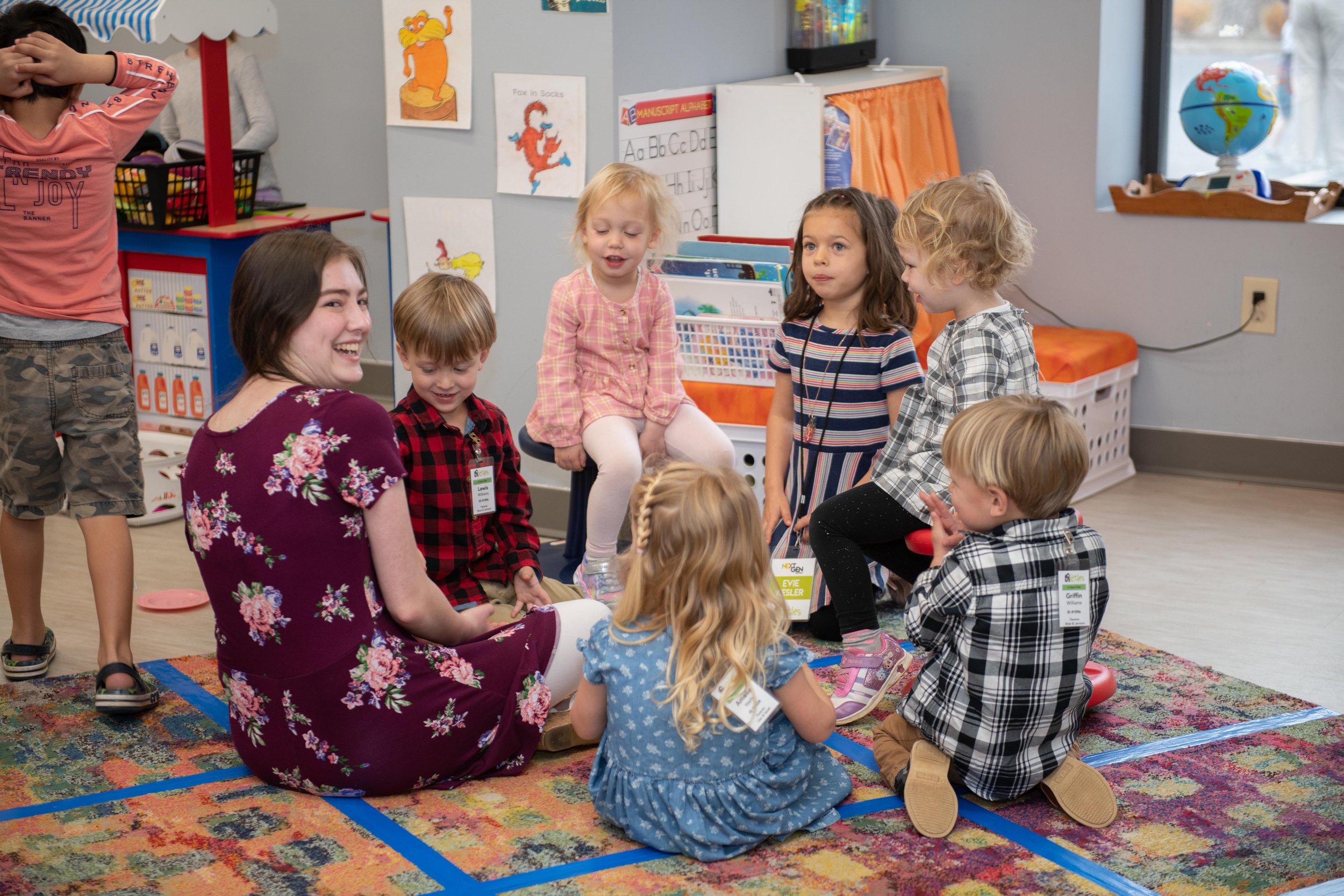 Young children and a teacher sitting on a colorful rug in a classroom, engaging in a group activity. The classroom is decorated with educational posters and toys, and a window shows outdoor scenery.