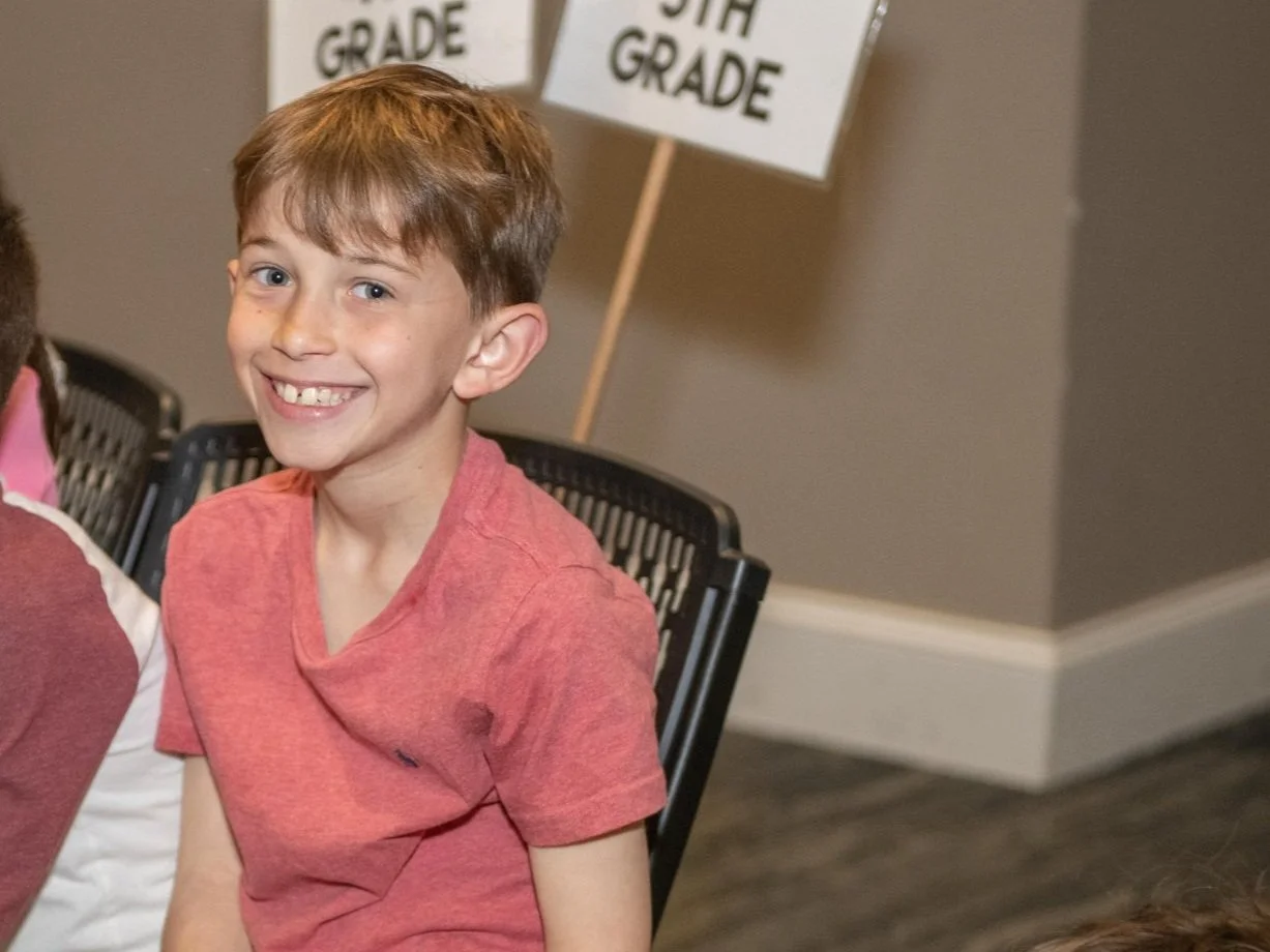 A young boy with light brown hair and a big smile sitting on a black chair in a room with gray walls and a sign behind him that reads '1st Grade'.