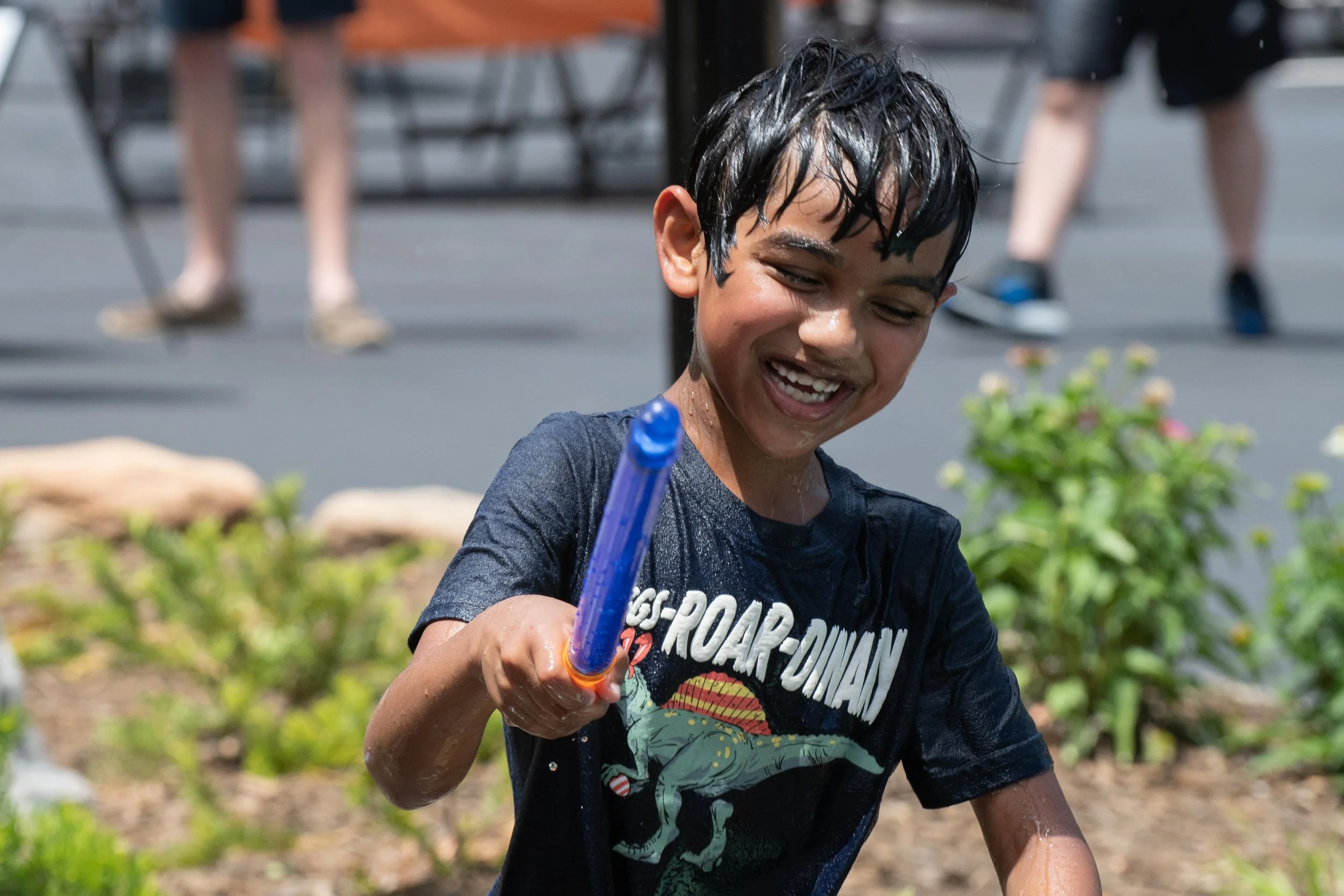 A young boy with wet black hair and a dark T-shirt smiling and playing outdoors with a water gun.