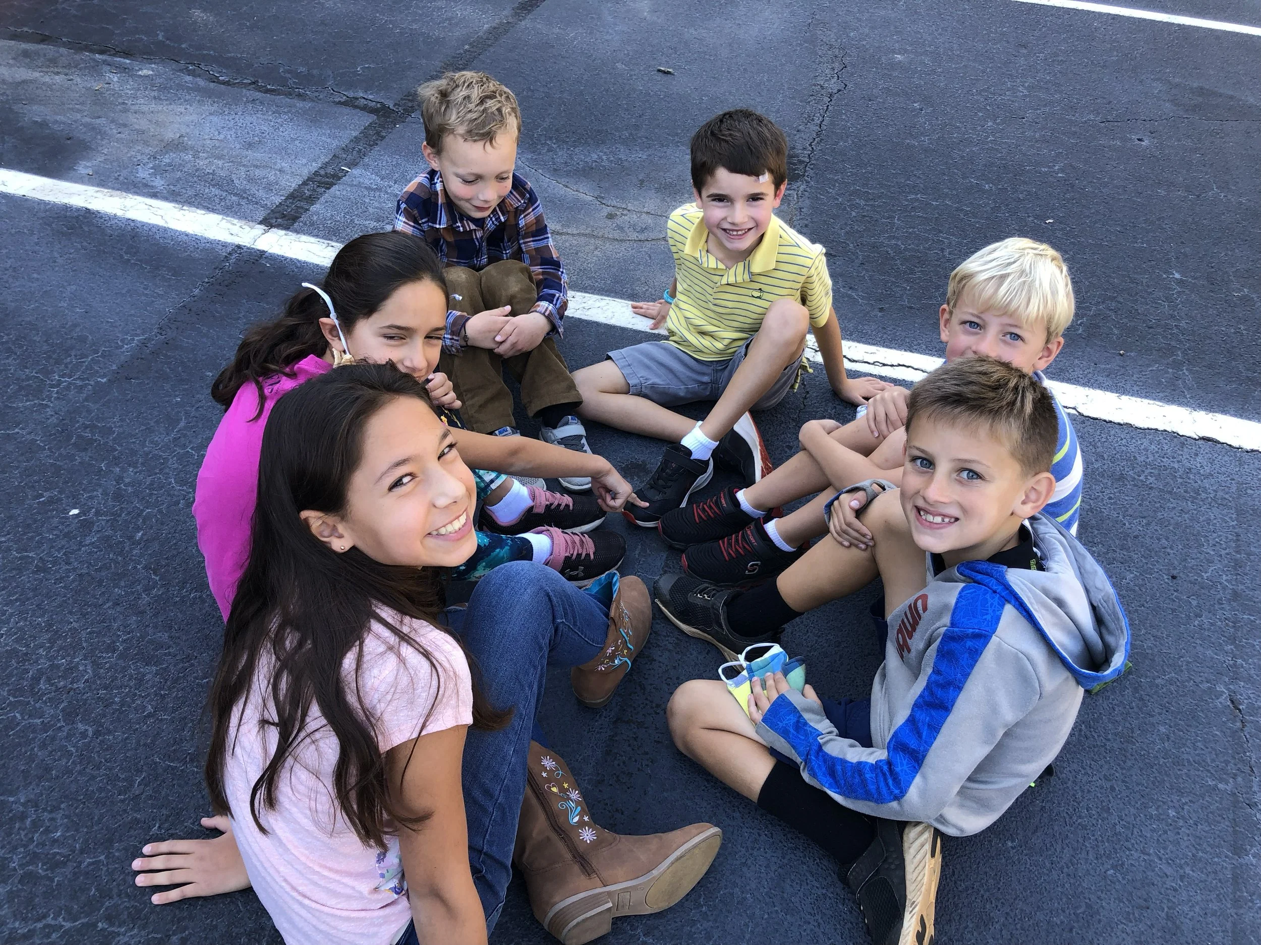Group of six children sitting on asphalt ground in a parking lot, smiling and looking at the camera.