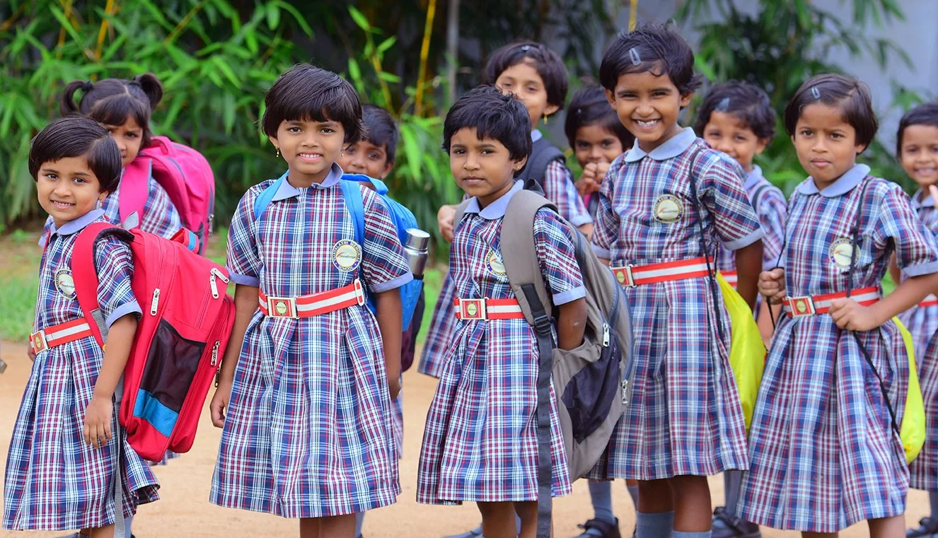 Group of young girls in school uniforms with backpacks standing outdoors, smiling and looking at the camera.