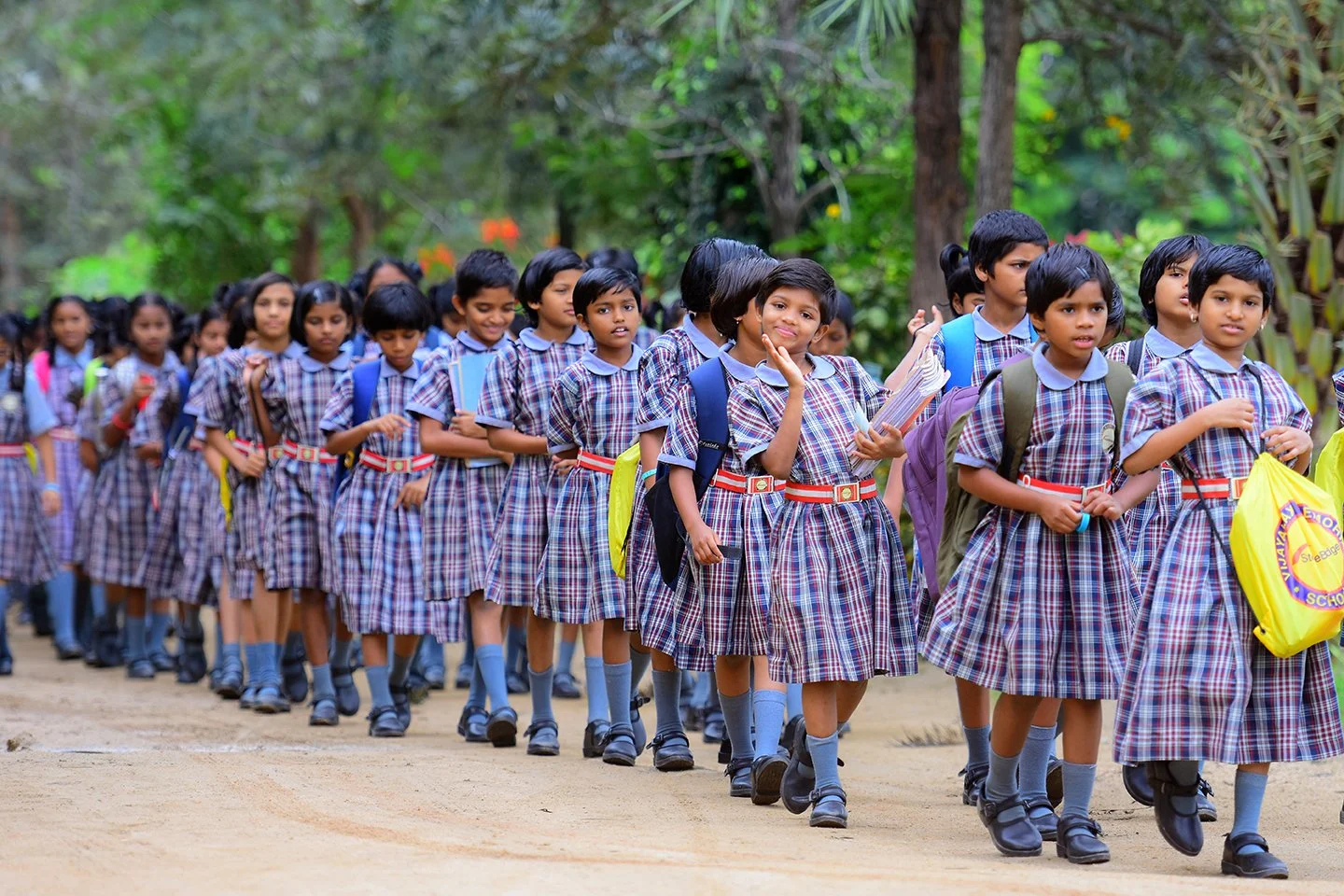 Group of school children in uniform walking outdoors on a dirt path surrounded by trees.