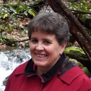 A woman smiling outdoors near a creek, surrounded by greenery and weathered trees.