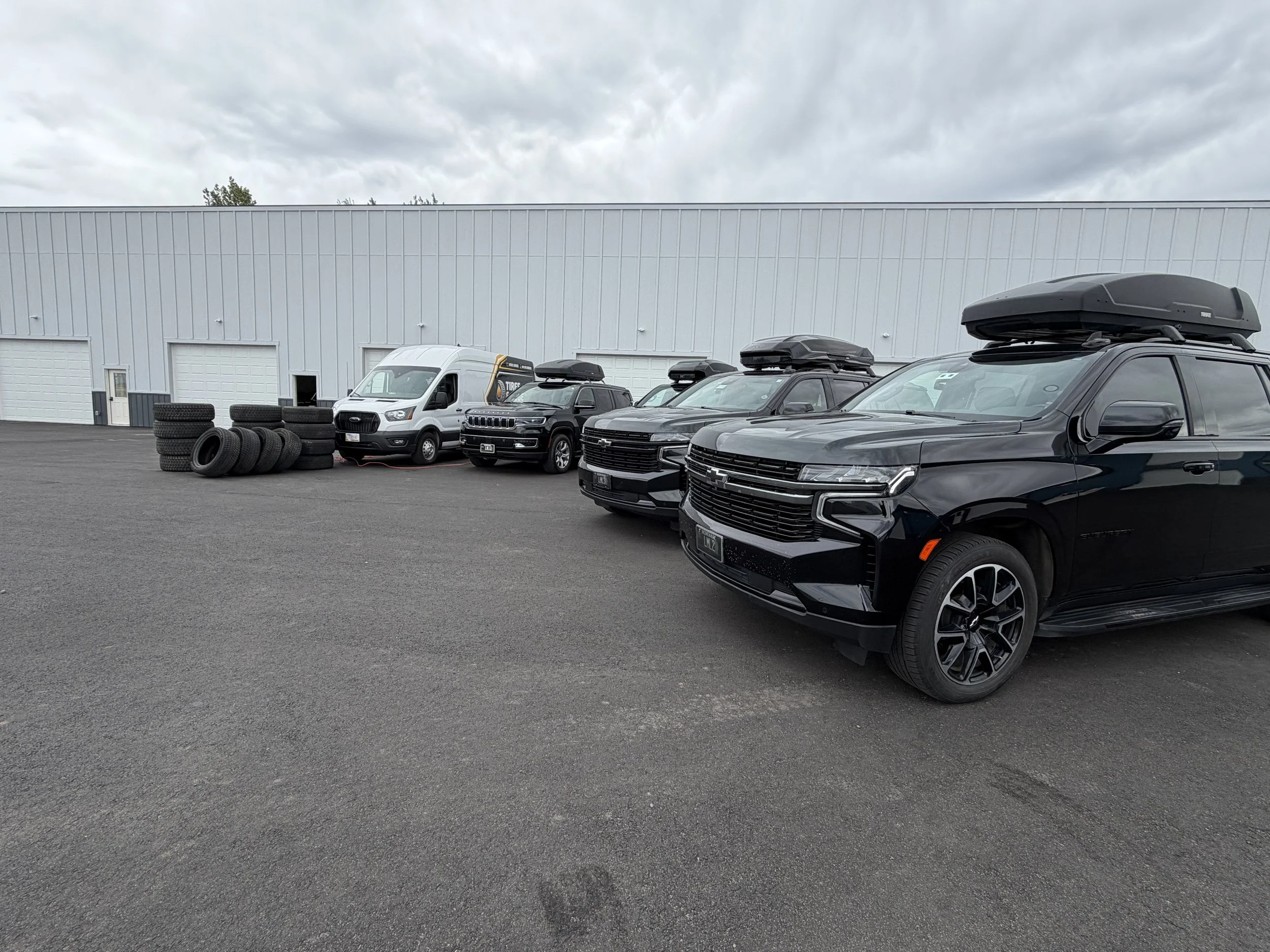 A row of parked cars and vans outside a gray industrial building with a large flat roof on a cloudy day.