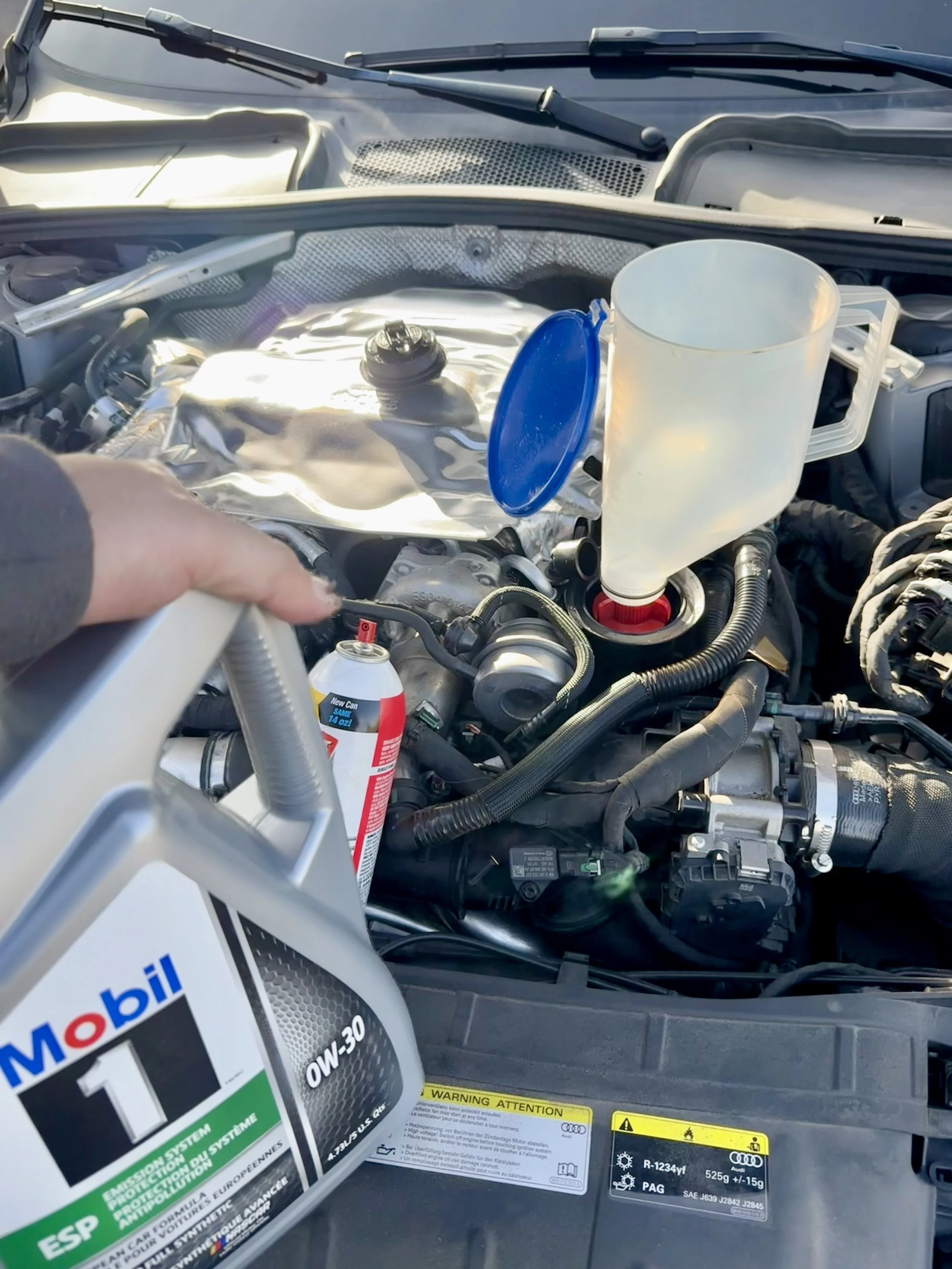 A car engine bay with a container of Mobil 1 synthetic motor oil in the foreground and the engine visible behind it.