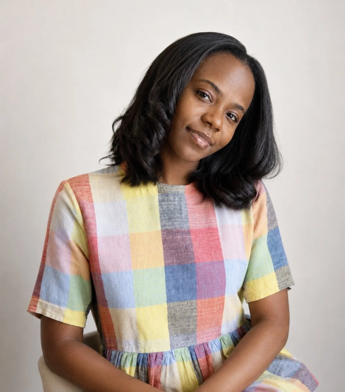 Portrait of a woman with shoulder-length black hair, wearing a colorful plaid dress, smiling softly against a plain light background.